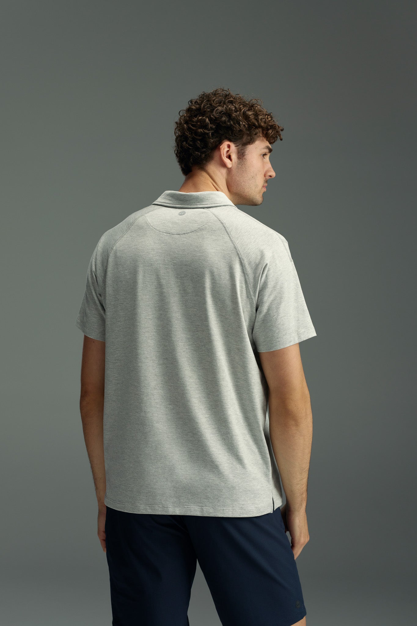 A man with curly hair is seen from behind wearing a WearEver Outbound Polo in light gray, made from breathable cotton-blend fabric, paired with dark shorts against a plain gray background.