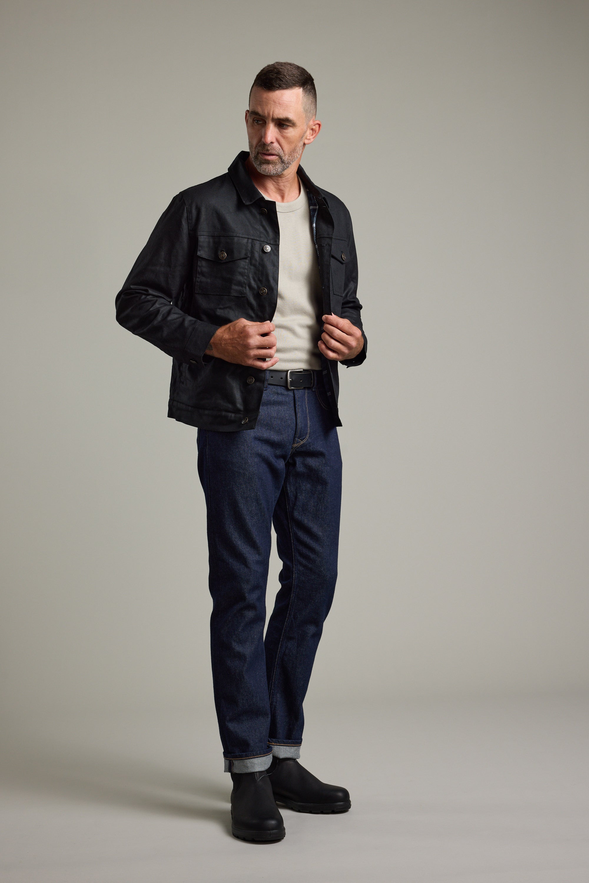 Man in a Barkers Benson Waxed Cotton Jacket, beige shirt, blue jeans, and black boots in studio.