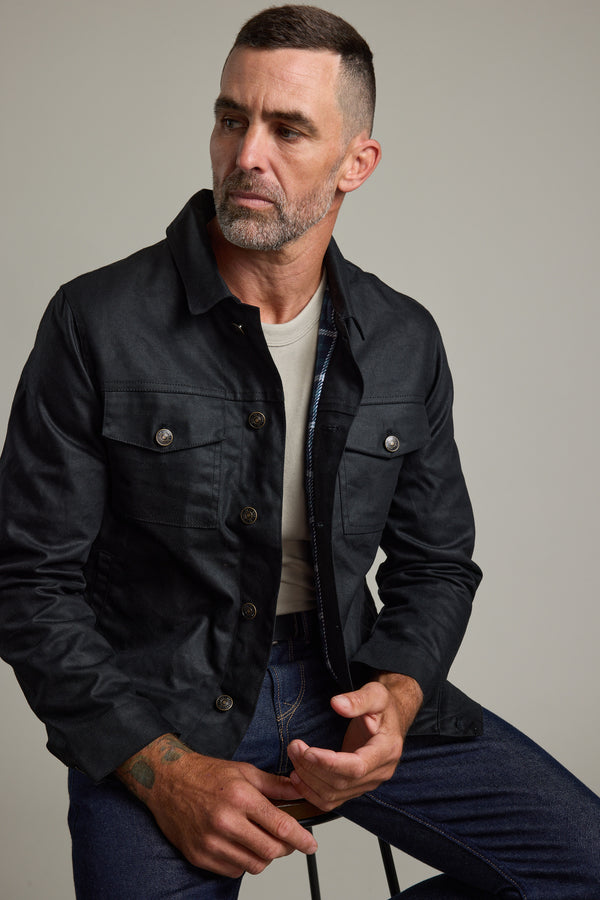 Man in Barkers Benson Waxed Cotton Jacket and jeans, sitting on a stool.