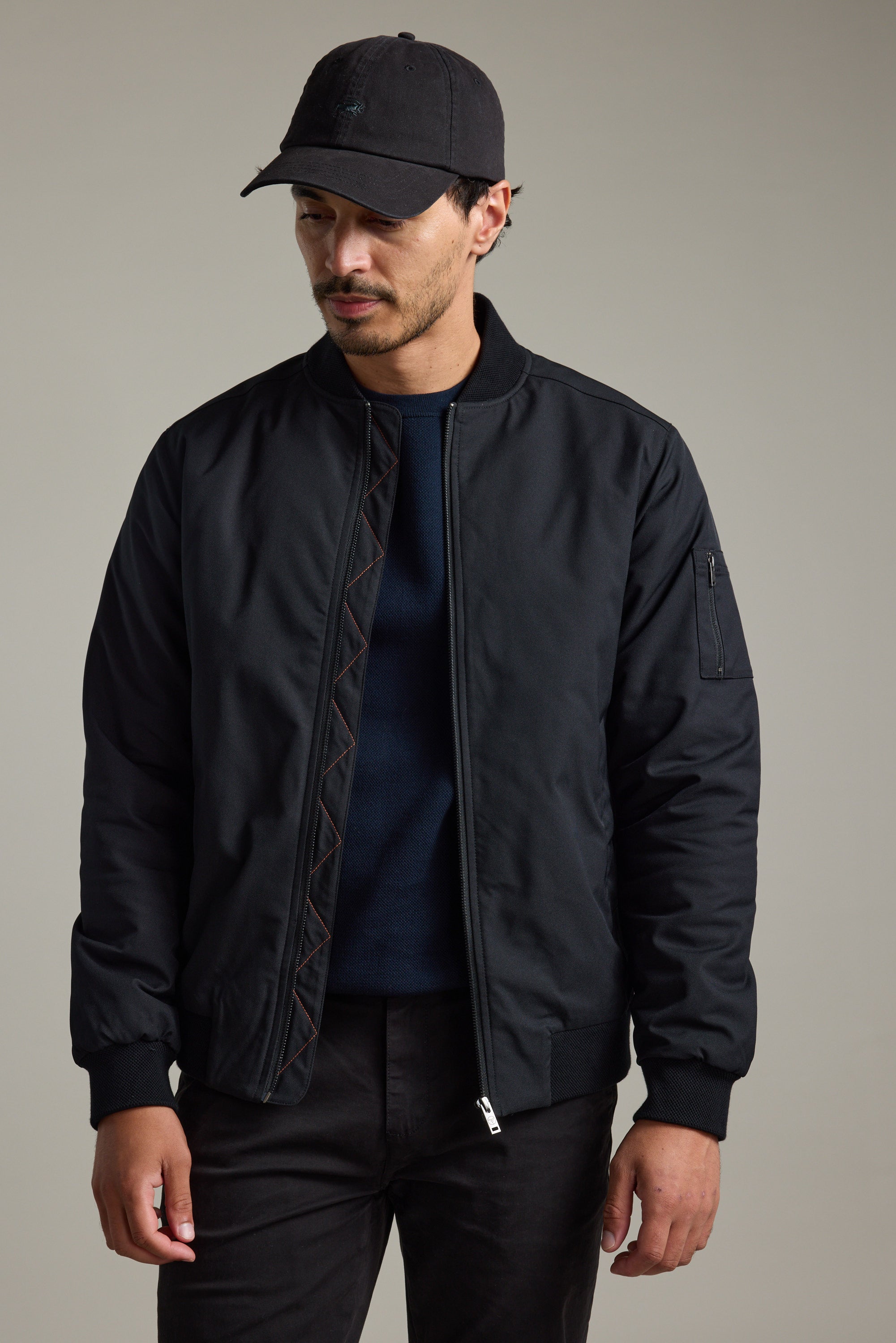 Man in a Barkers Dunhill Bomber Jacket, black pants, and cap stands against a plain background.