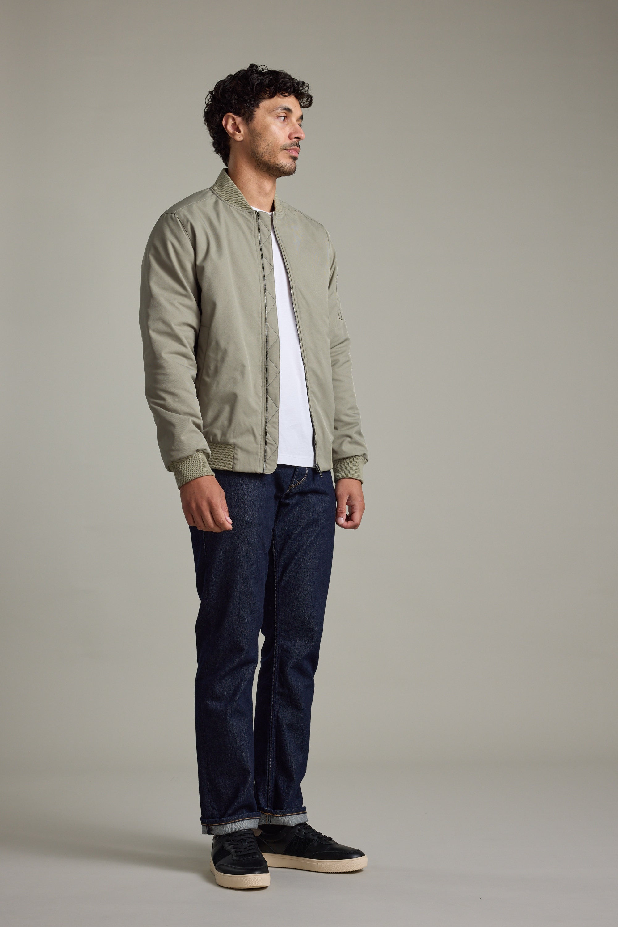 Man in a Barkers Dunhill Bomber Jacket, white shirt, jeans, and sneakers on plain background.