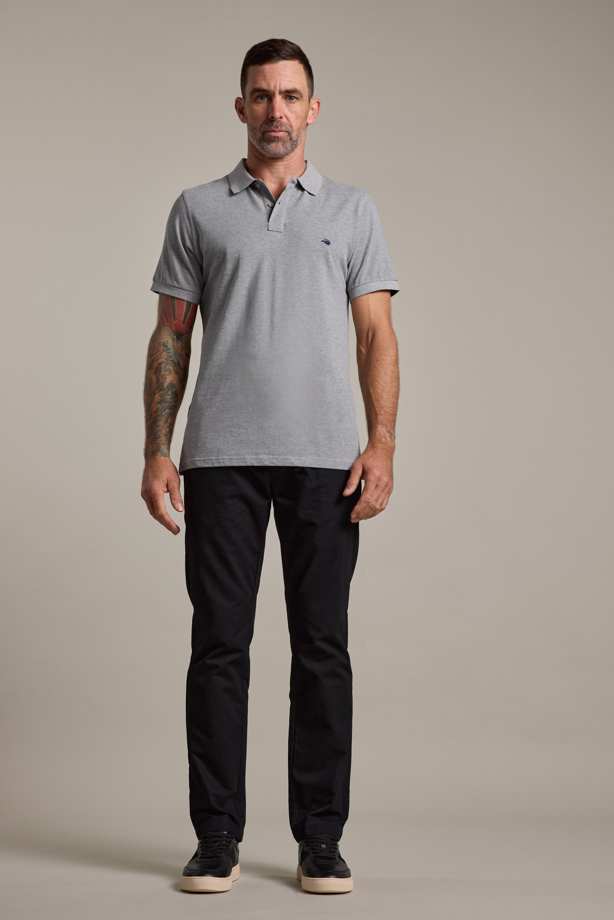 A man with short dark hair and a tattooed left arm stands facing forward, wearing a light gray polo shirt, Barkers Dundas Chino trousers in relaxed fit cotton twill, and black sneakers against a plain beige background.