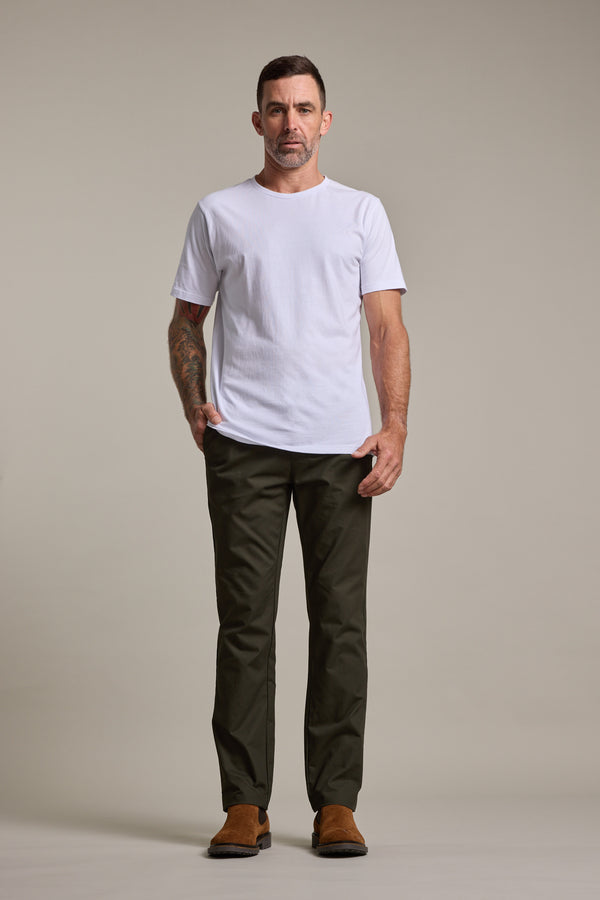 A man stands facing forward with one hand in his pocket, wearing a plain white t-shirt, Barkers Dundas Chino trousers in dark green relaxed fit, and brown shoes against a plain beige background.