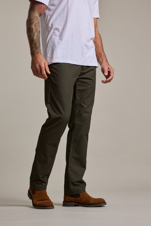 A person stands against a plain background, showing only the lower half in a white t-shirt, Barkers Dundas Chino trousers in olive green, and brown suede boots. One visible arm is decorated with a colorful tattoo.