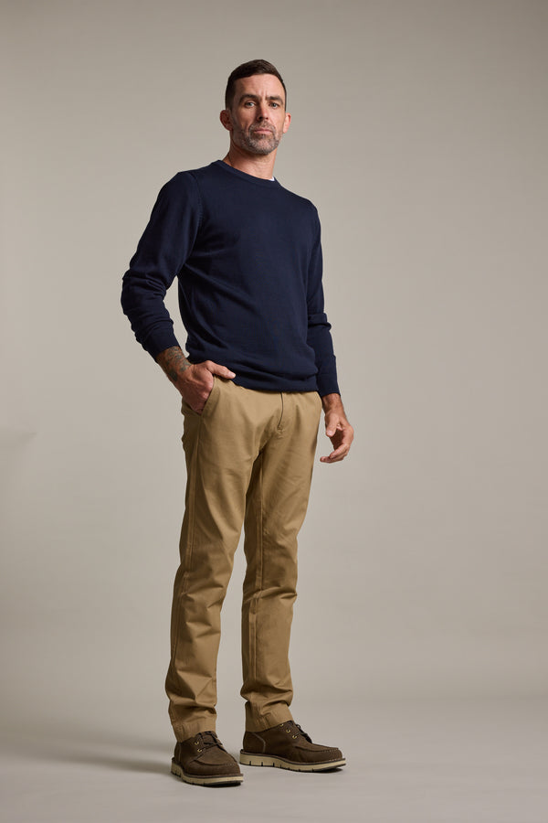A man with short dark hair, a trimmed beard, and neutral expression stands against a plain background, wearing a navy sweater, Barkers Dundas Chino relaxed fit trousers in lightweight cotton twill, and brown shoes with one hand in his pocket.