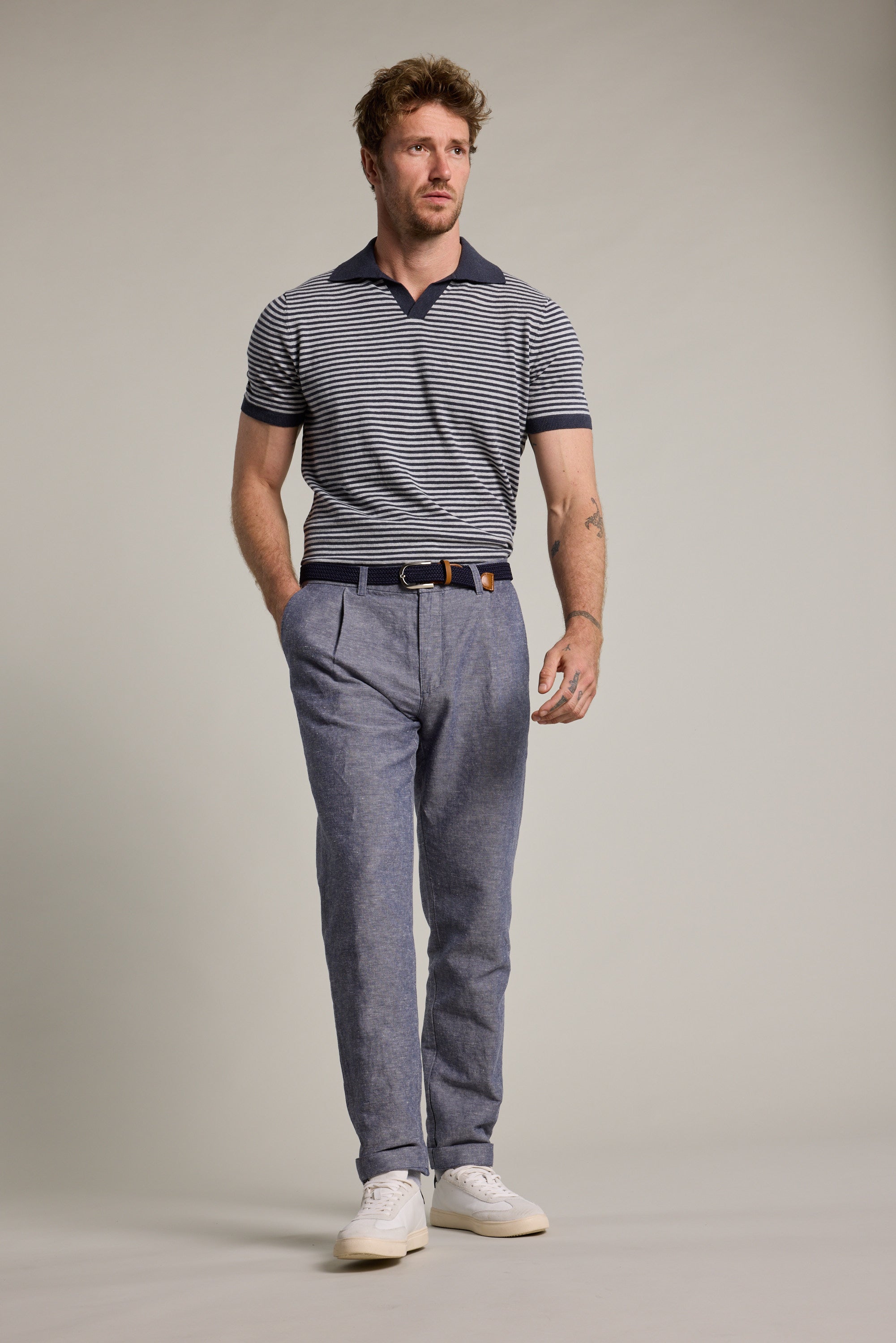 A man with short brown hair and a beard wears a navy striped polo, Barkers Newman Linen Pants, a dark belt, and white sneakers, standing against a plain light gray background.