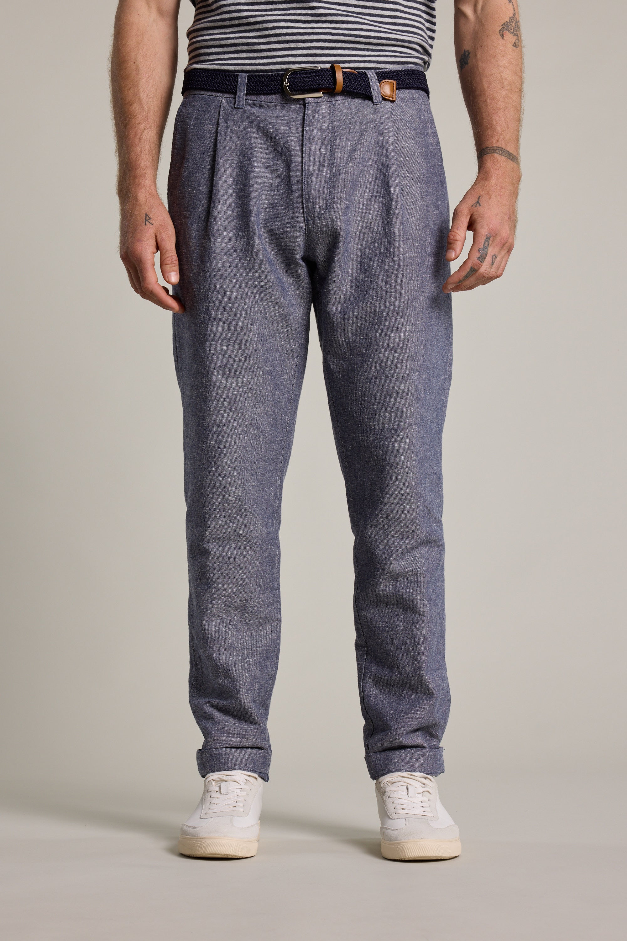 A person, visible from shoulders down, wears Barkers Newman Linen Pants in blue with a dark belt, a tucked-in striped shirt, and white sneakers, standing against a plain light background.