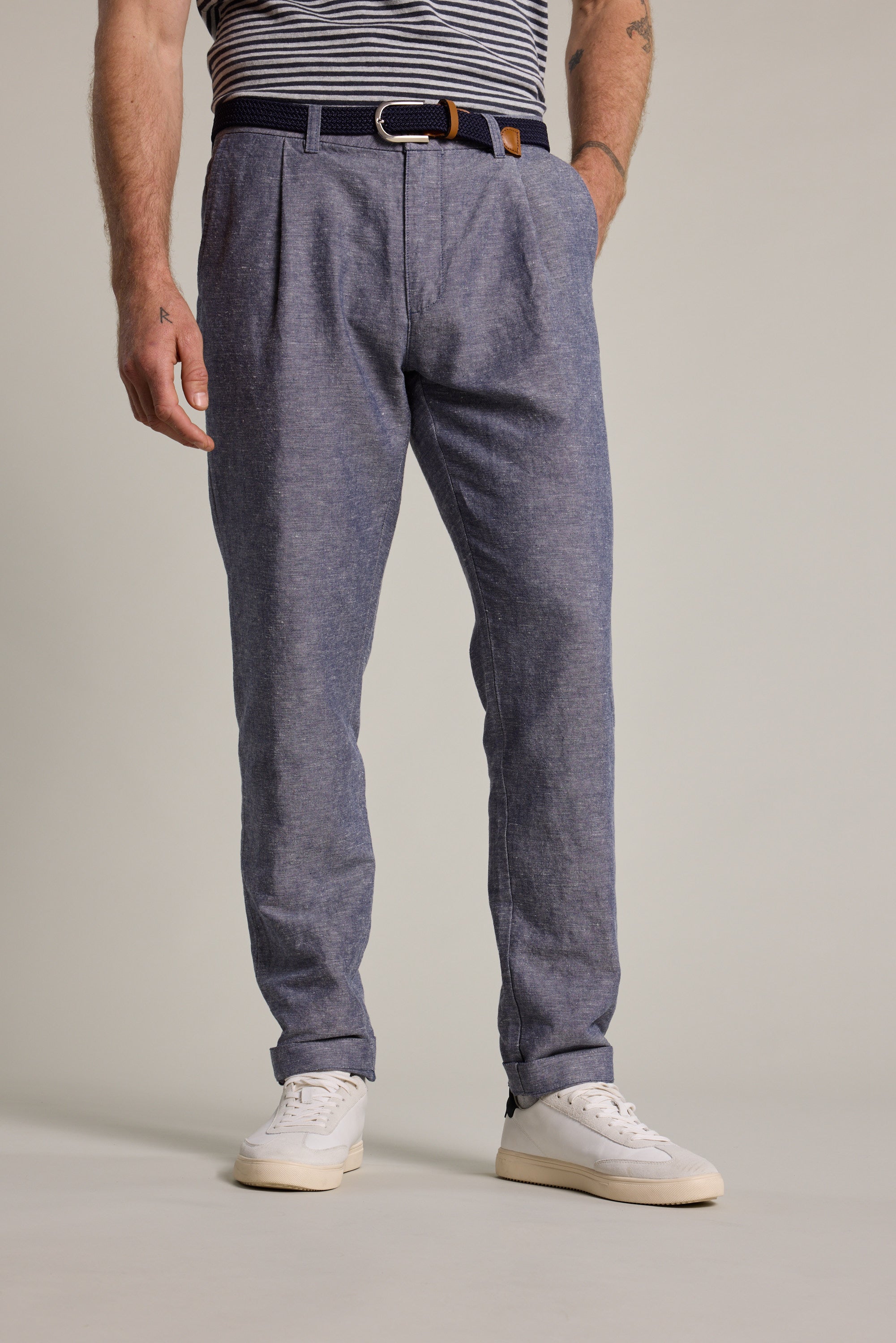 A man stands against a plain light background, hands in pockets, wearing Barkers' Newman Linen Pants with a striped shirt tucked in, black belt, and white sneakers.