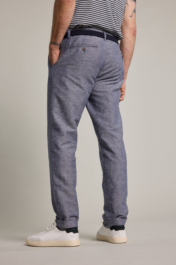 A person faces away from the camera against a plain light background, hands in pockets, wearing Barkers Newman Linen Pants with a striped shirt and white sneakers.