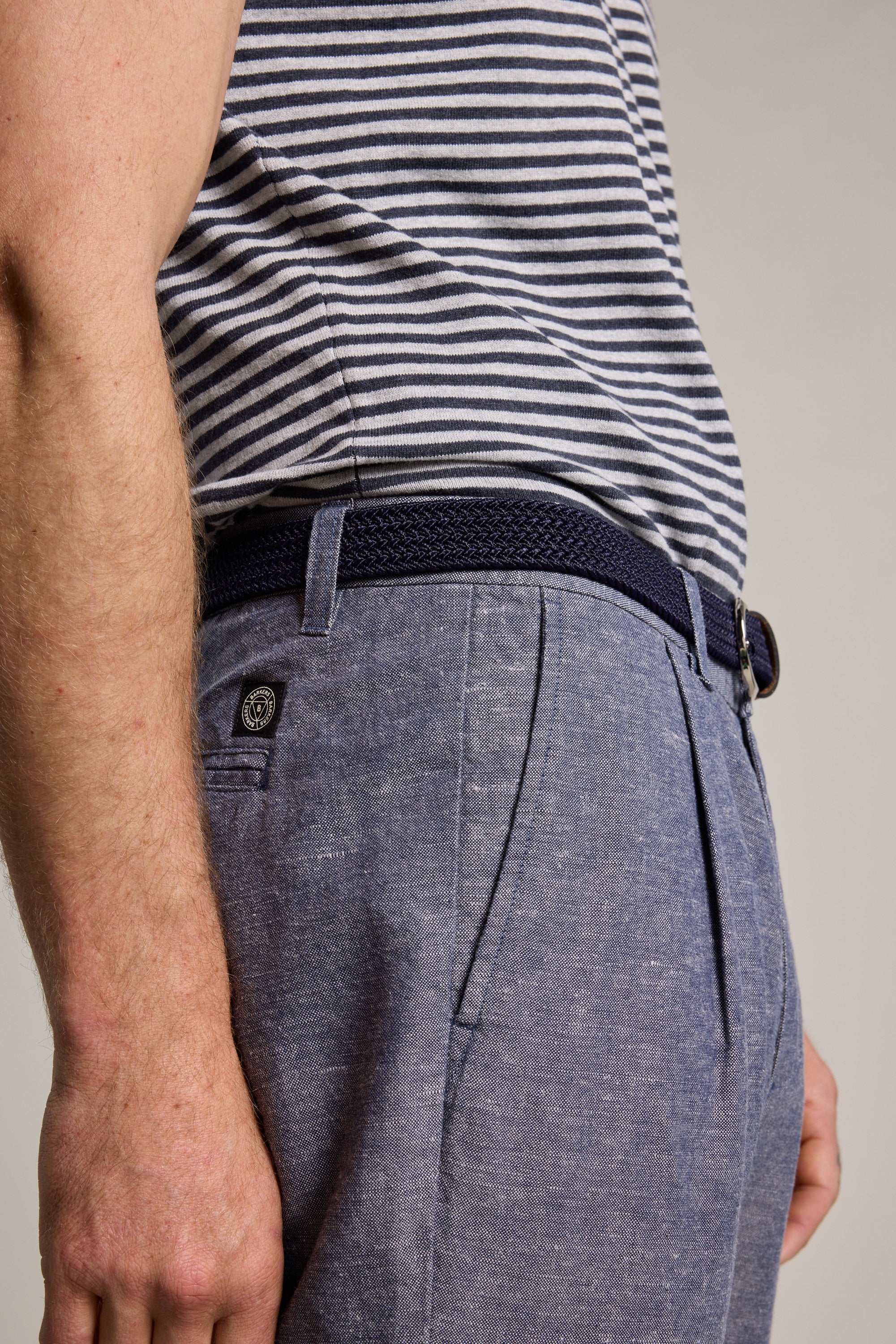 A person wears blue Barkers Newman Linen Pants with a gray and white striped short-sleeve shirt and navy braided belt, seen from the side at waist height for a smart-casual look.