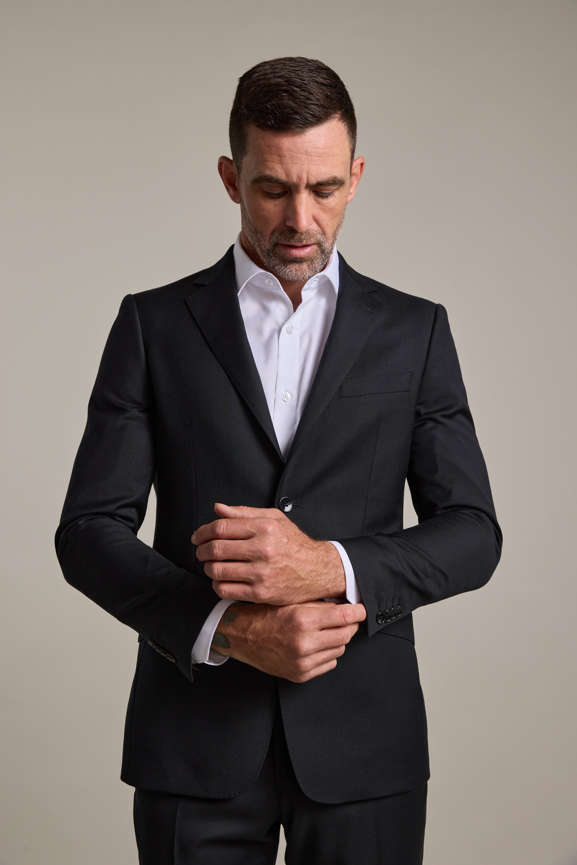 A man wearing the Barkers Mayfair ZQrx Suit Jacket and a white dress shirt stands against a plain background, looking down as he adjusts his left cuff with his right hand.
