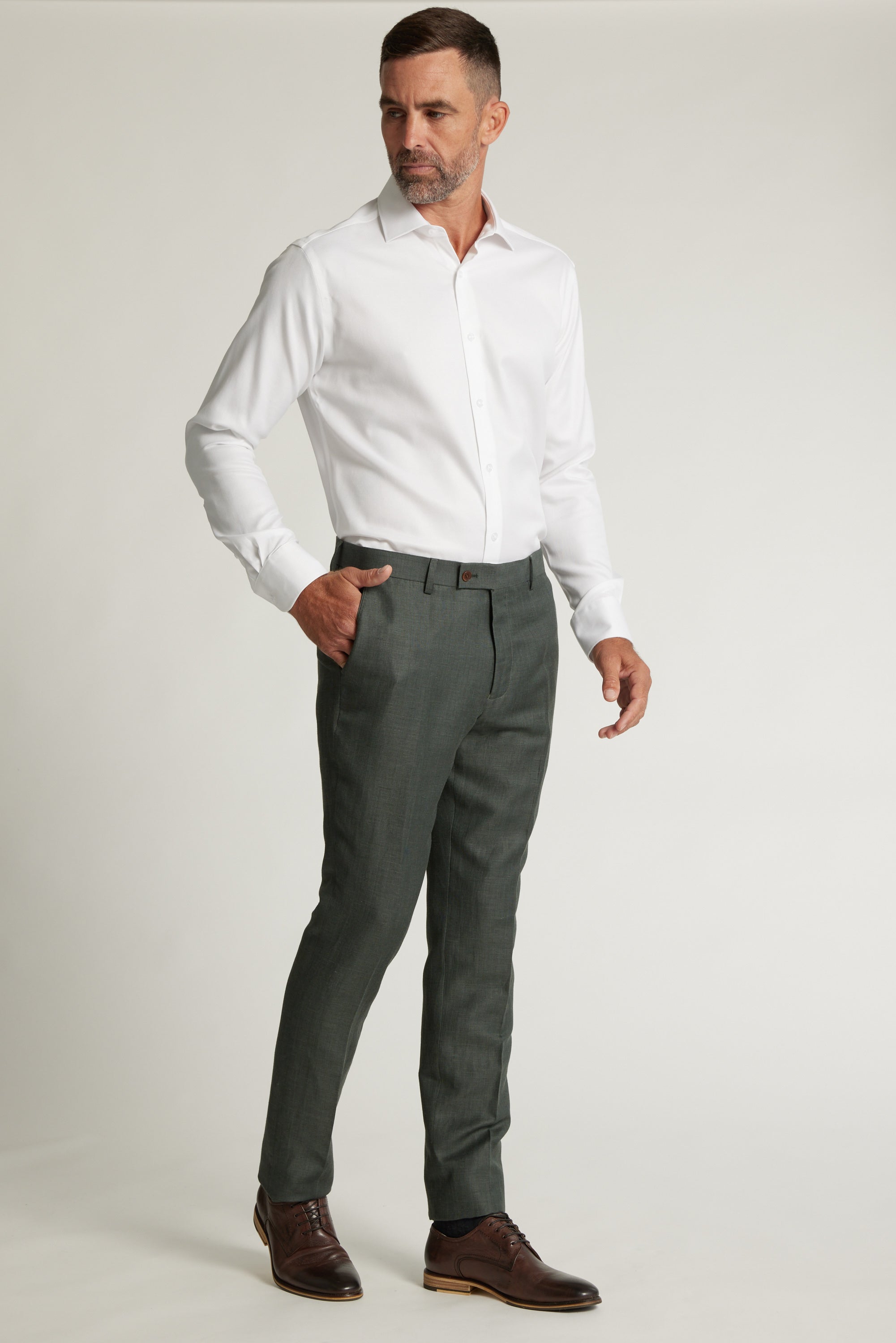 A man with short hair and a beard stands against a plain background, wearing a white shirt tucked into gray Barkers Inverness Suit Trousers with a belt and brown dress shoes, one hand in his pocket.