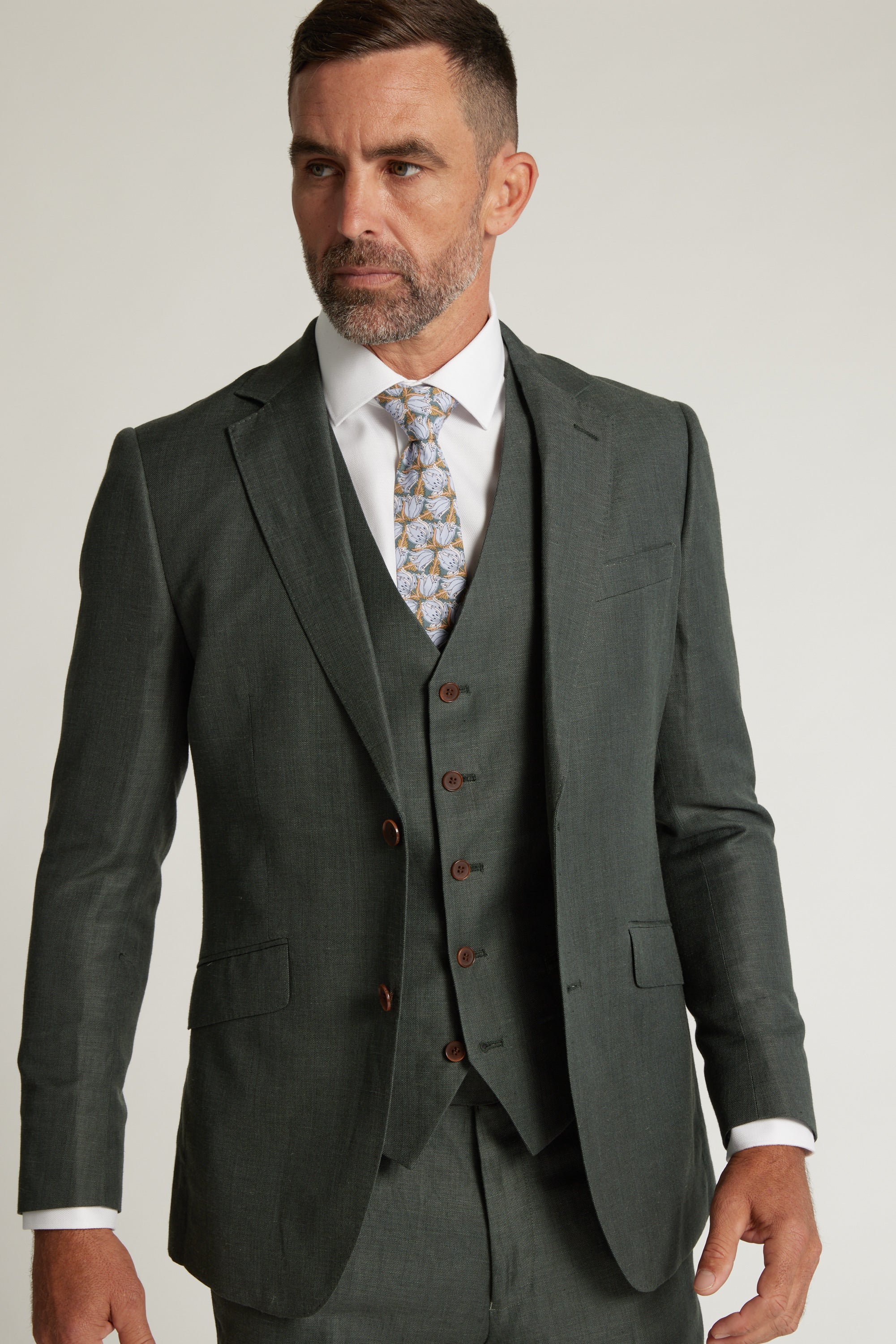 A man with short brown hair and a beard wears the Barkers Inverness Suit Jacket in sage green with a white dress shirt and patterned tie, looking to the side against a plain light background.