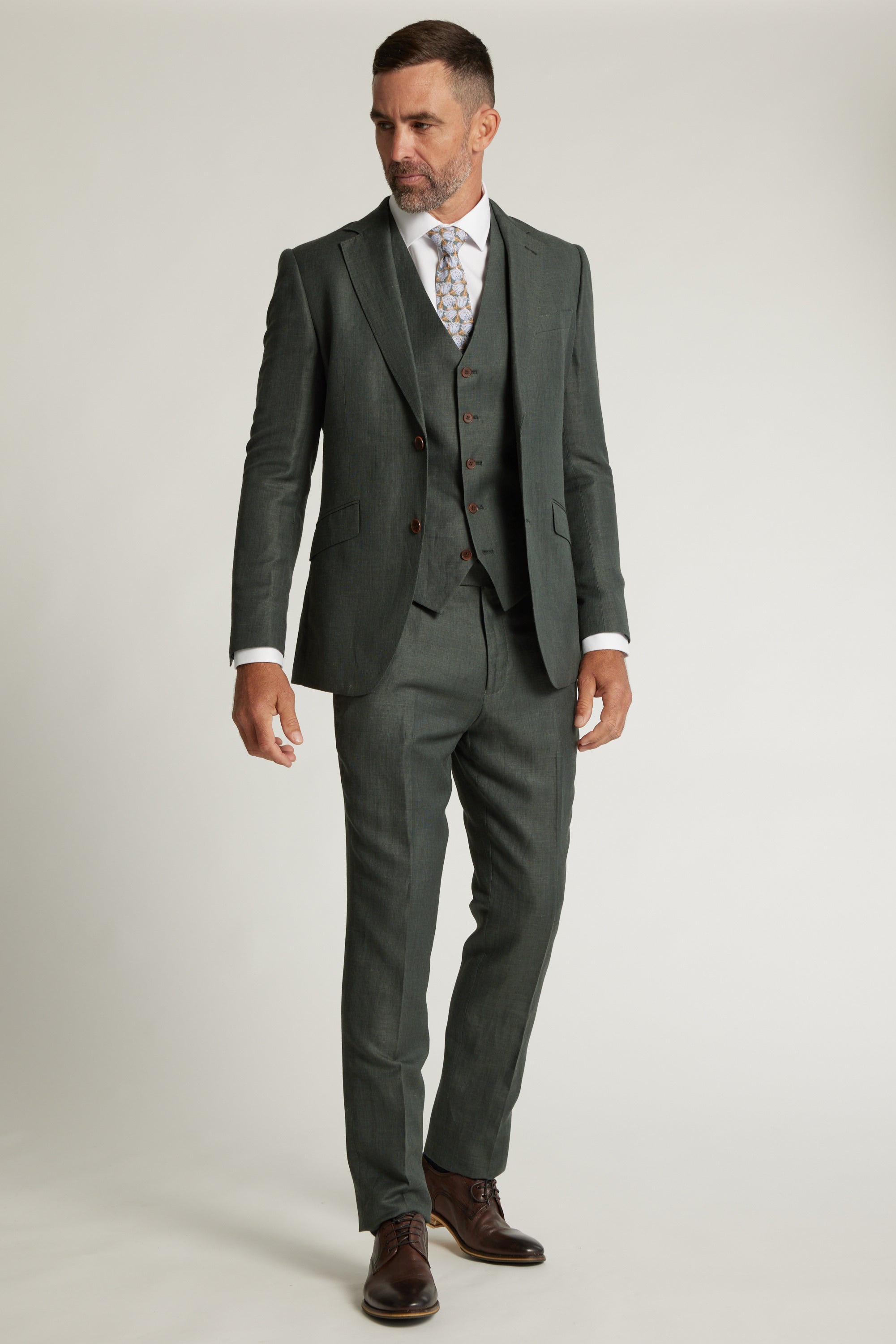 A man wears the Barkers Inverness Suit Jacket in sage green with a white shirt, patterned tie, and brown dress shoes, standing against a plain light background.