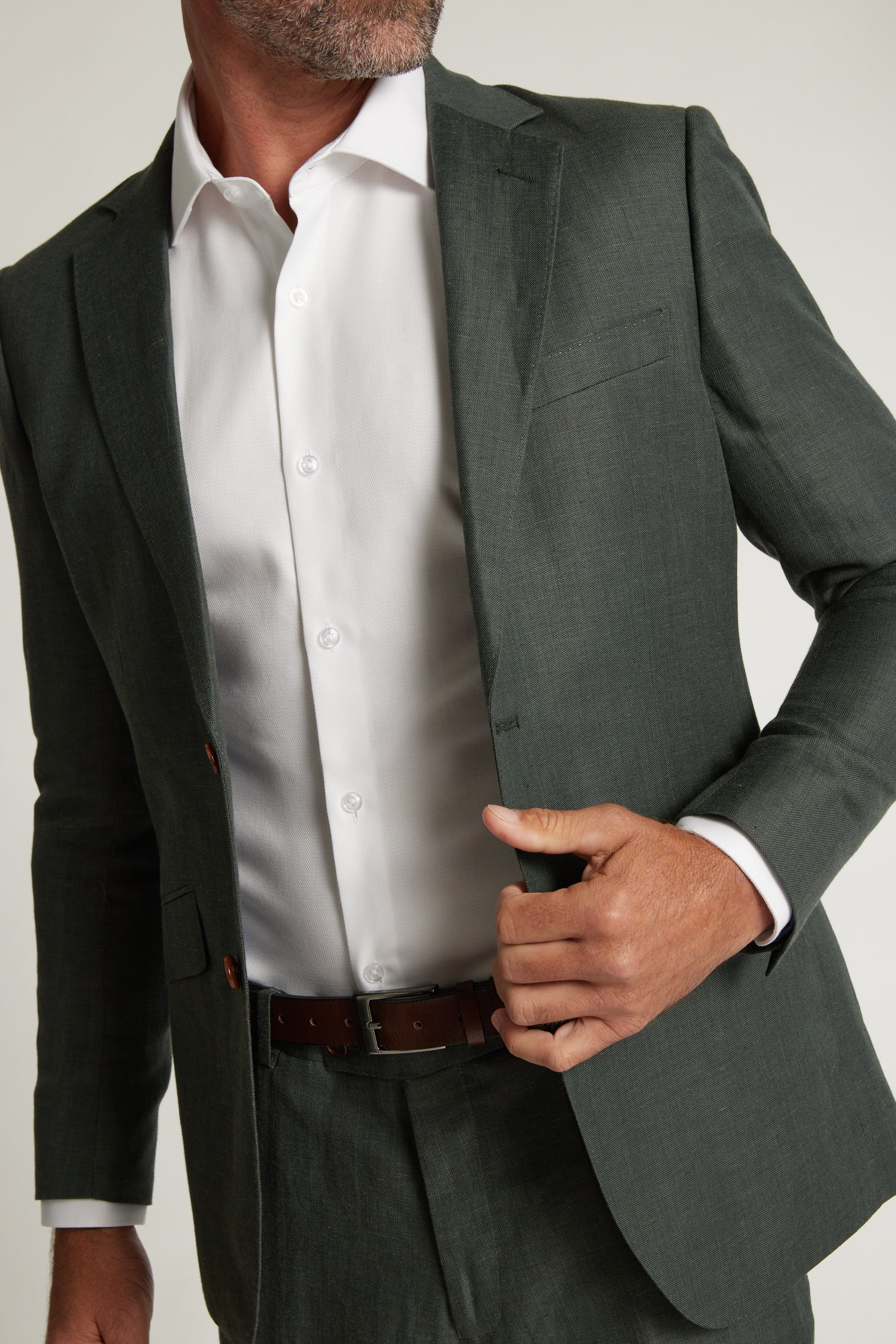 A man in a Barkers Inverness Suit Jacket in sage green over a white dress shirt adjusts his jacket with one hand, his face partly out of frame.