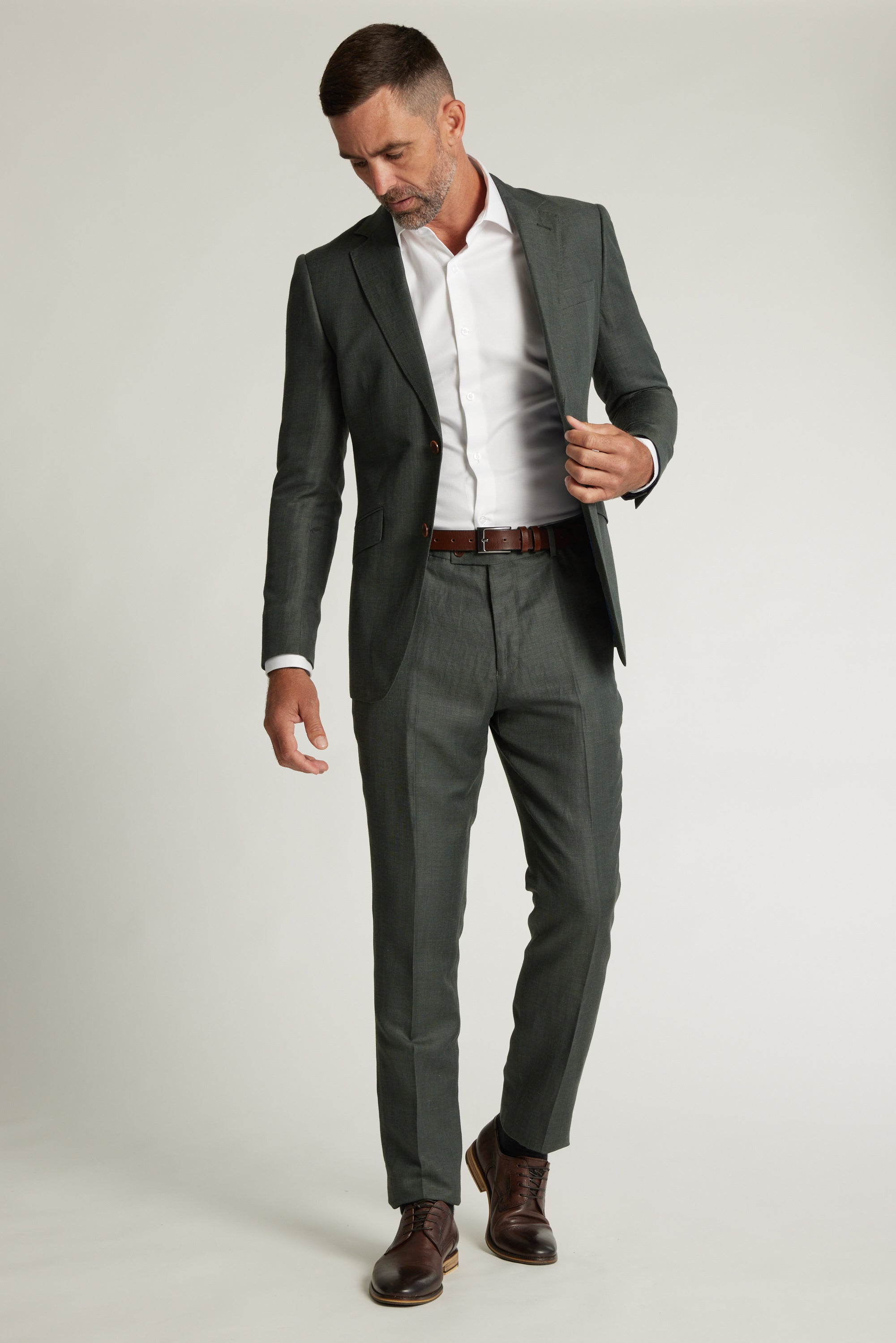 A man in a fitted sage green Inverness Suit Jacket by Barkers, paired with a white dress shirt, brown belt, and brown dress shoes stands against a plain light background, looking down as he adjusts his jacket with one hand.