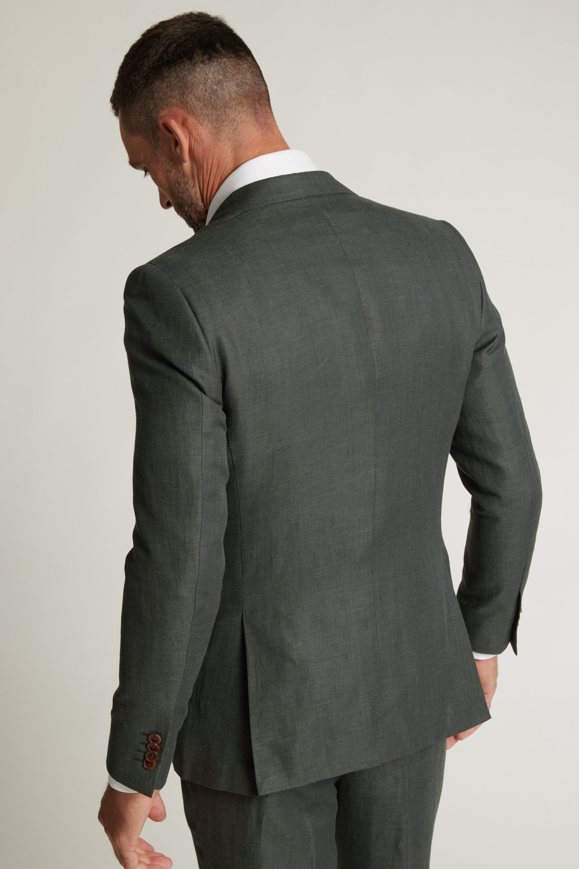 A man in a Barkers Inverness Suit Jacket in sage green stands with his back to the camera and head slightly lowered, set against a plain light background.