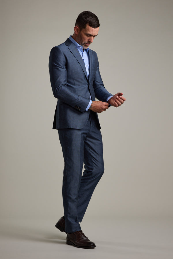 A man wearing Barkers' Charles Texture Suit Trouser in steel grey and a light blue dress shirt stands against a plain background, looking down as he adjusts his sleeve. He wears dark dress shoes and has short hair.