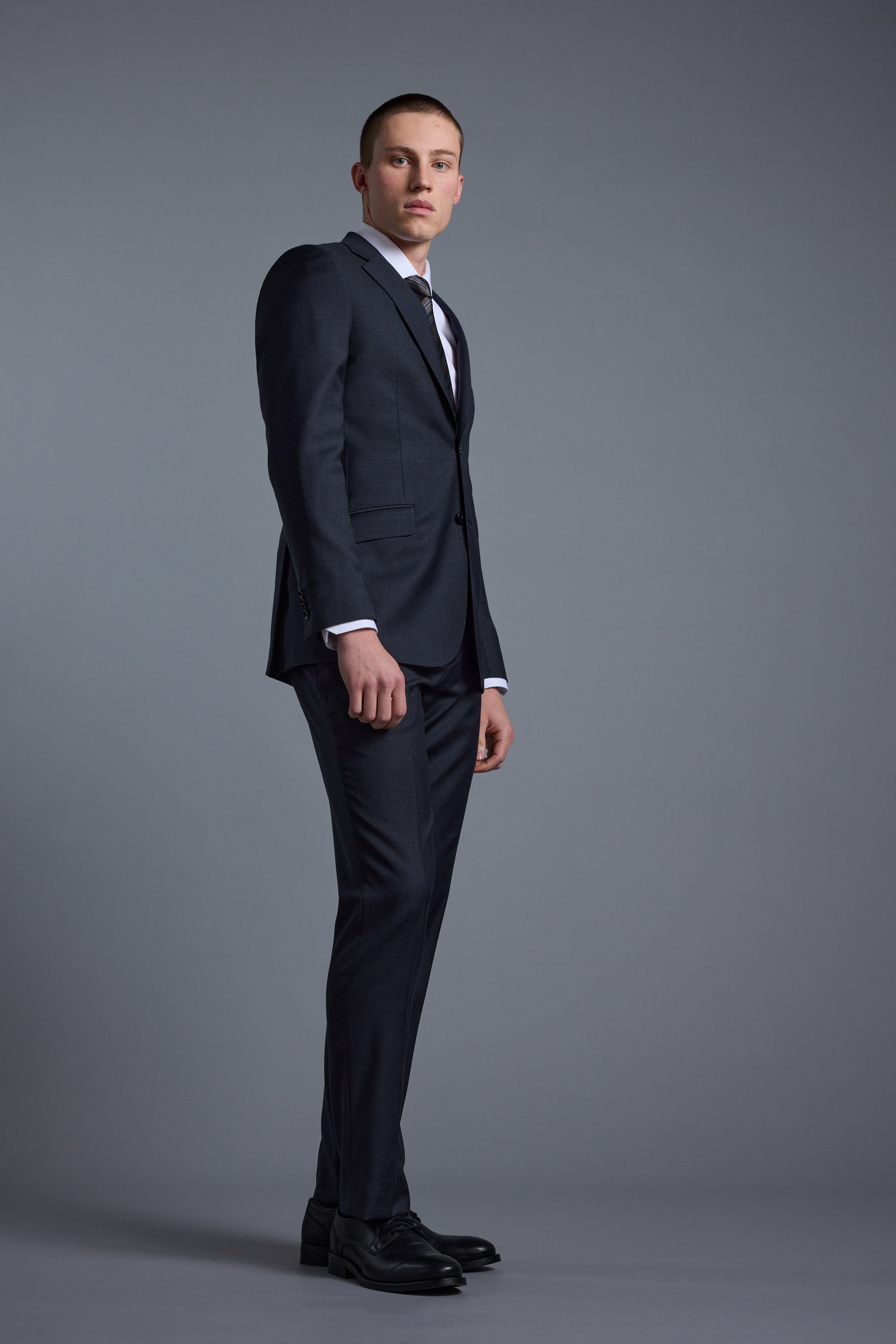 A young man in Barkers’ Mayfair ZQrx Suit Trouser, paired with a dark suit jacket, white shirt, and tie, stands against a plain gray background, gazing slightly left with a neutral expression.