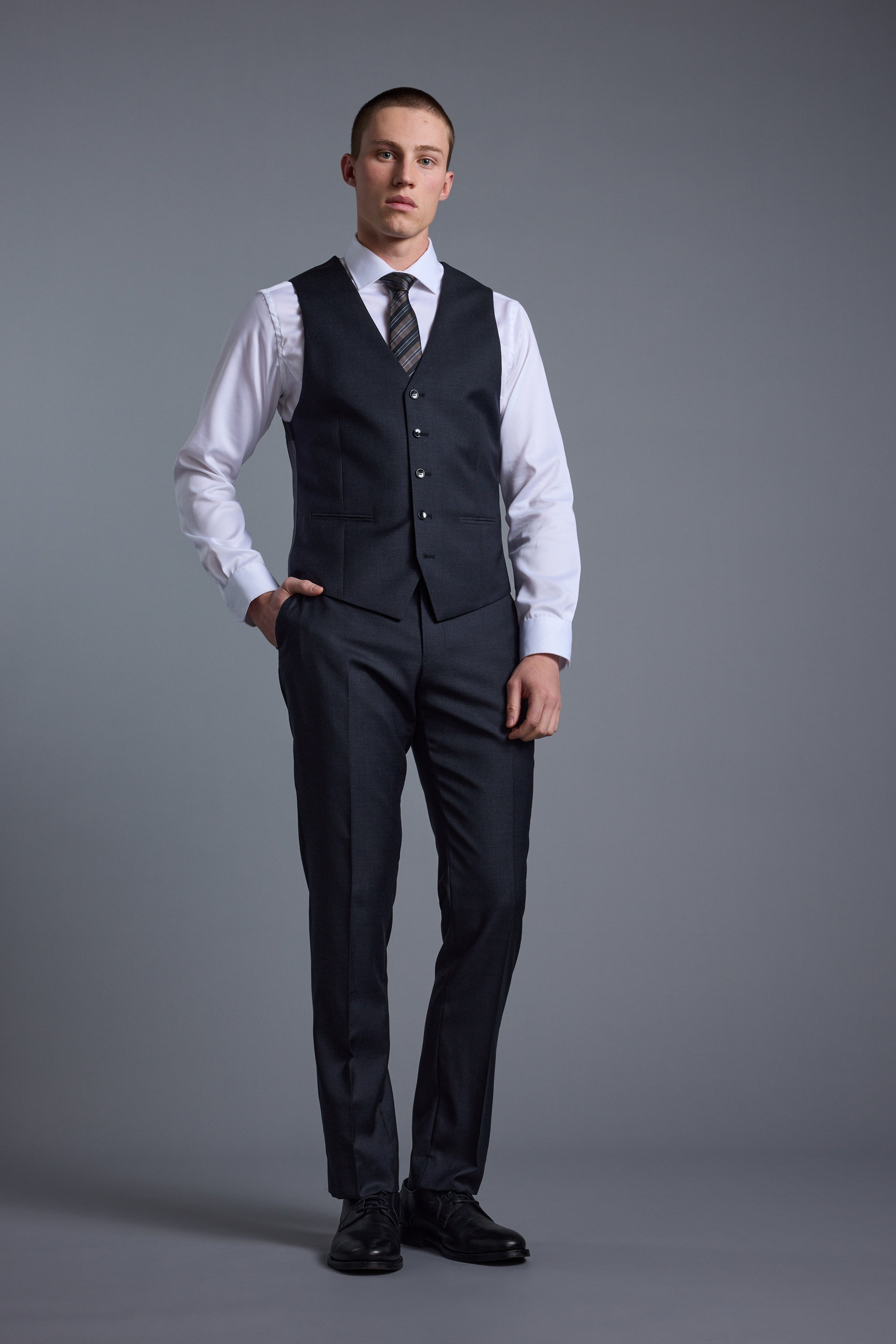 A man stands against a gray background wearing Barkers Mayfair ZQrx Suit Trousers with a matching dark jacket, white dress shirt, patterned tie, and black dress shoes, one hand in his pocket and a neutral expression.