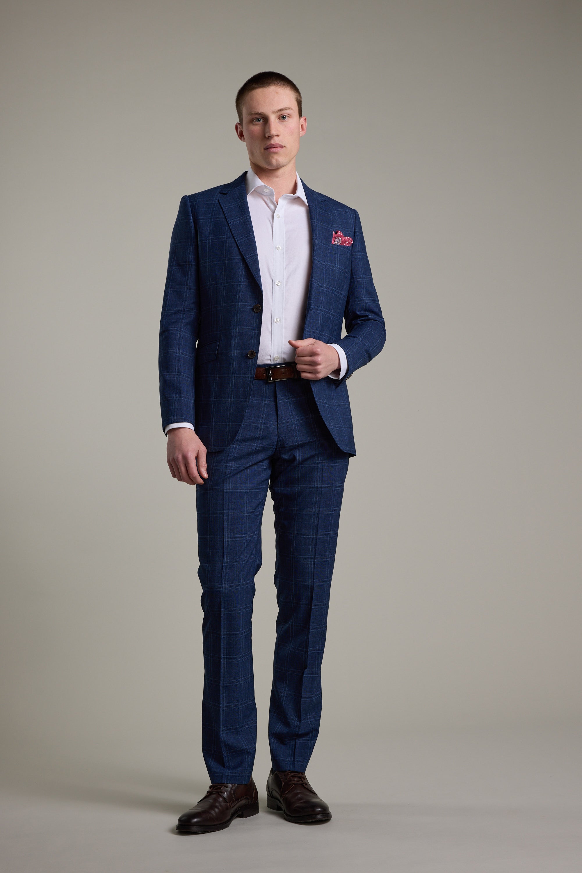 A man stands against a plain background, wearing the Barkers Leonard Check Suit Jacket with a white dress shirt, brown belt, brown shoes, and a red pocket square, looking straight at the camera with a neutral expression.