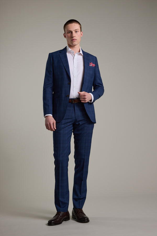 A man stands against a plain background, wearing the Barkers Leonard Check Suit Jacket with a white dress shirt, brown belt, brown shoes, and a red pocket square, looking straight at the camera with a neutral expression.