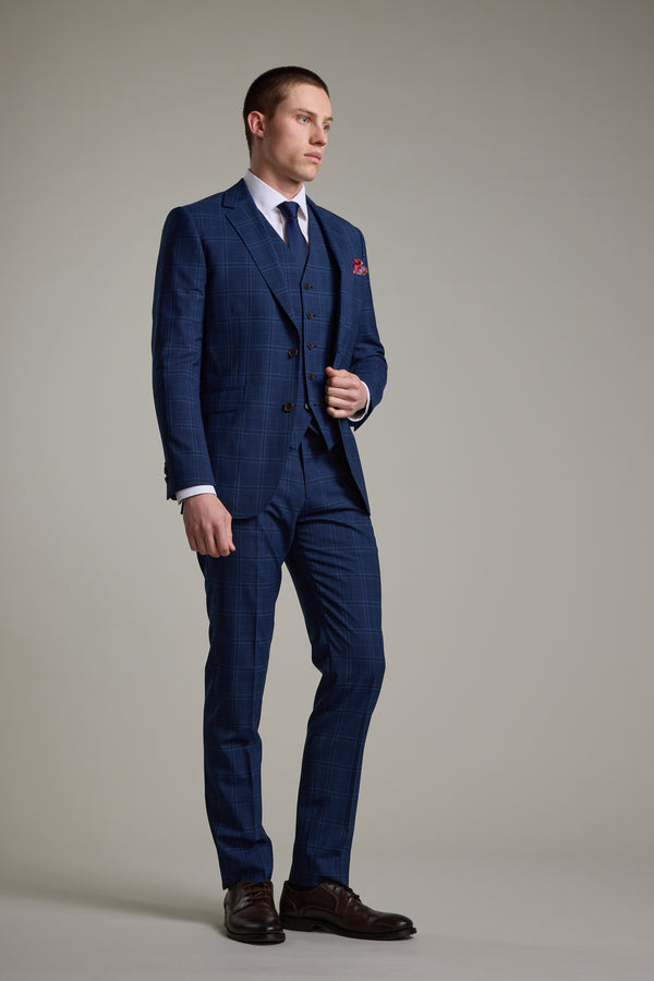 A man stands against a plain background wearing the Barkers Leonard Check Suit Jacket, styled with a white shirt, dark tie, brown shoes, and a red pocket square, with one hand adjusting his jacket.