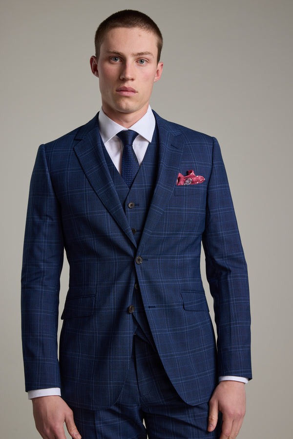 A young man in the Barkers Leonard Check Suit Jacket, a navy windowpane three-piece, with a white dress shirt, dark tie, and red patterned pocket square stands against a light background, looking at the camera with a neutral expression.