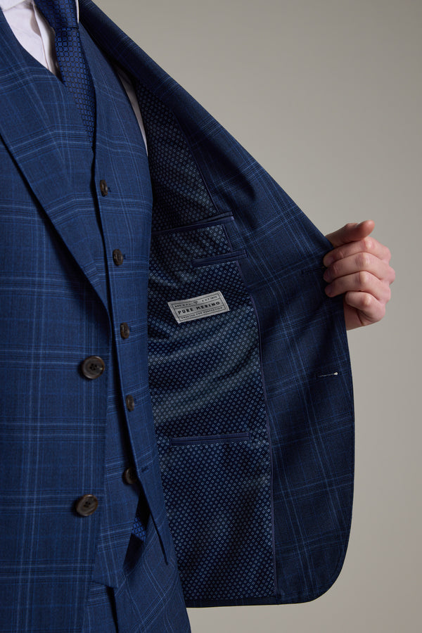 A person wears the Barkers Leonard Check Suit Jacket in navy windowpane Merino wool, holding the jacket open to show its patterned blue lining and interior label, with their hand visible near the side seam.