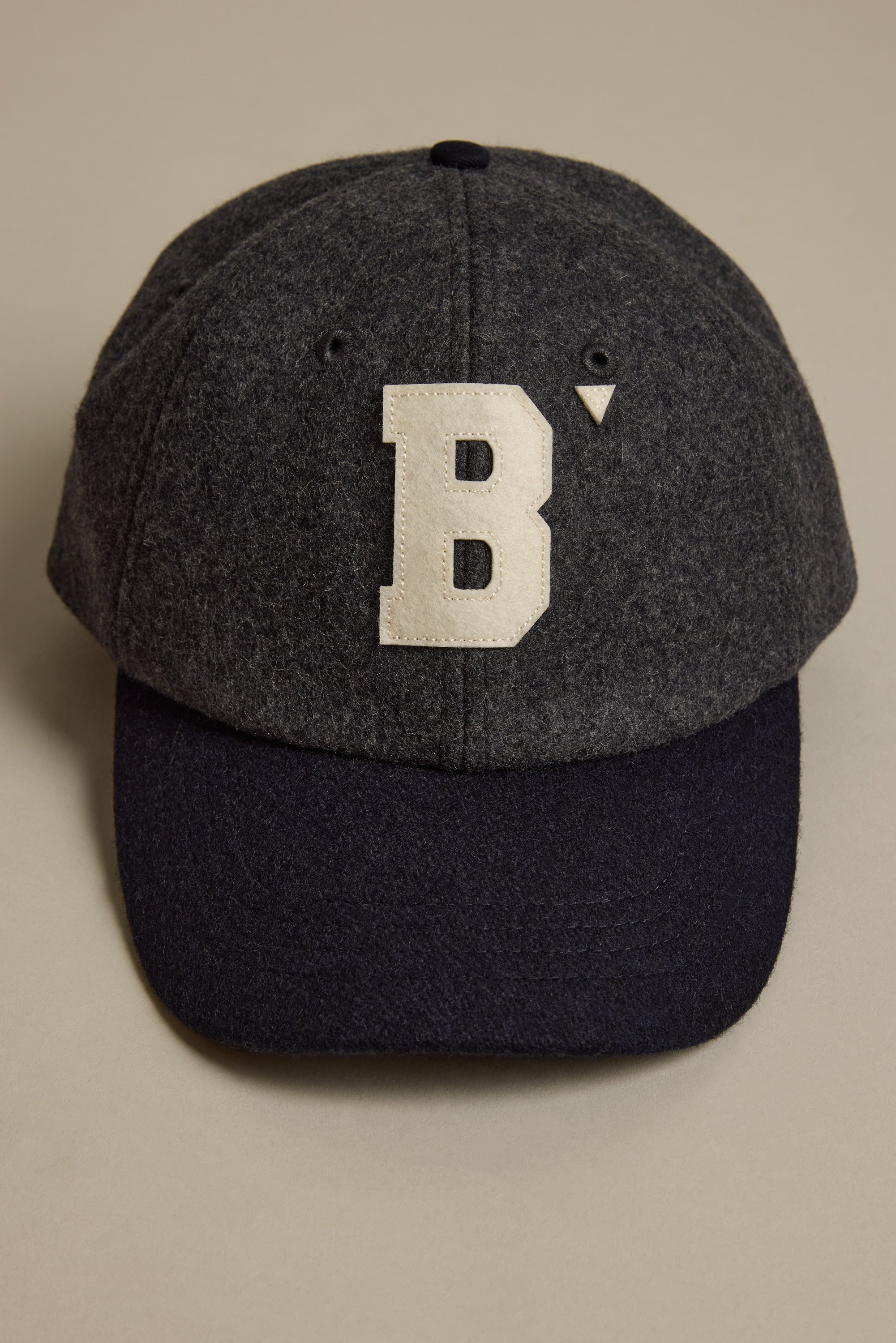 The Darcy Woollen Cap by Barkers is a dark gray wool cap with a black brim and features the Barkers B logo—an oversized white B with a small triangle—on the front. The adjustable cap is displayed on a beige surface.