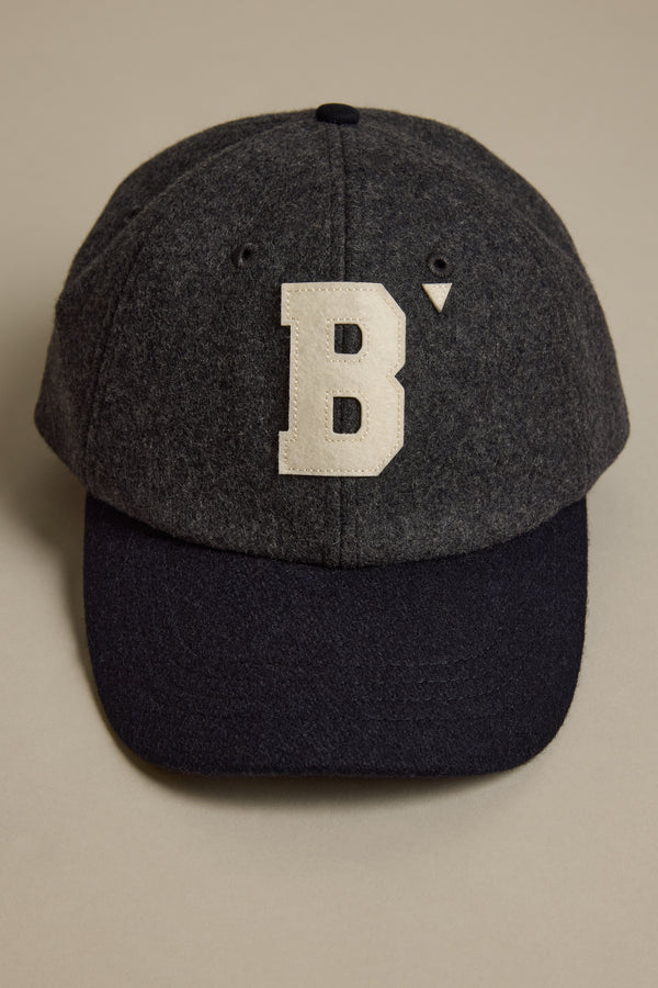 The Darcy Woollen Cap by Barkers is a dark gray wool cap with a black brim and features the Barkers B logo—an oversized white B with a small triangle—on the front. The adjustable cap is displayed on a beige surface.