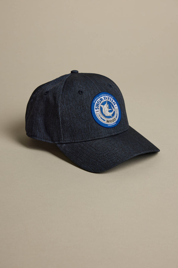 The Swordfish Mojo Trucker Cap by Swordfish Mojo is a dark blue denim cap with a round front patch featuring a swordfish illustration and the text “Swordfish Mojo,” shown on a neutral beige background.