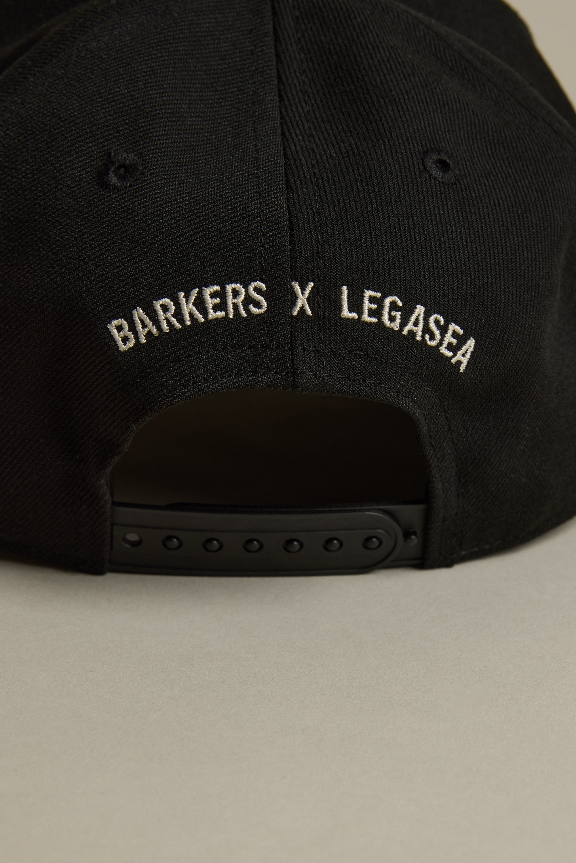 Close-up of the back of the black Legasea Logo Snapback Cap with an adjustable strap. White embroidered Legasea and BARKERS X LEGASEA logos appear above the strap for a customizable fit.