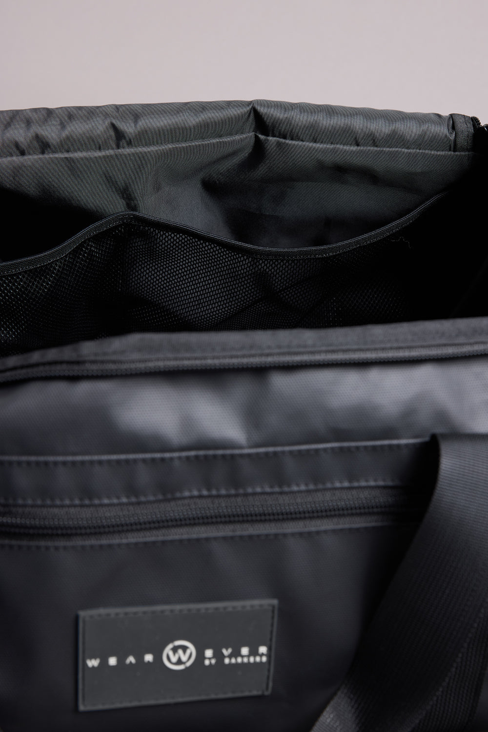 Close-up of the WearEver Transit Holdall’s open interior shows mesh compartments and a sneaker pocket. The black, water-resistant exterior has a zip pocket and displays the WearEver logo on a circular label.