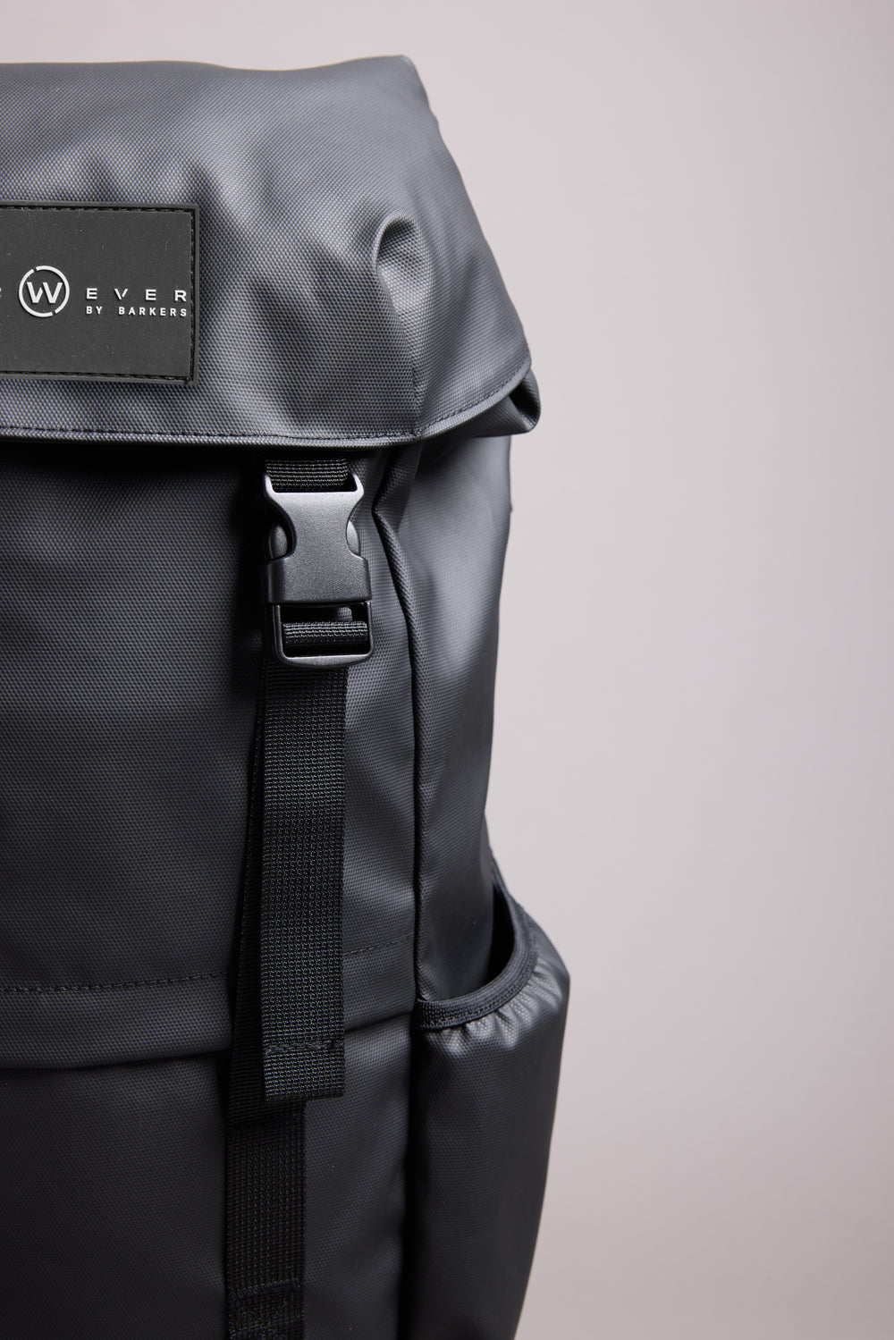 Close-up of the sleek black WearEver Transit Backpack, featuring a flap cover, water-resistant zips, black buckle strap, and a small "WearEver" logo patch on the upper left side against a plain light gray background.