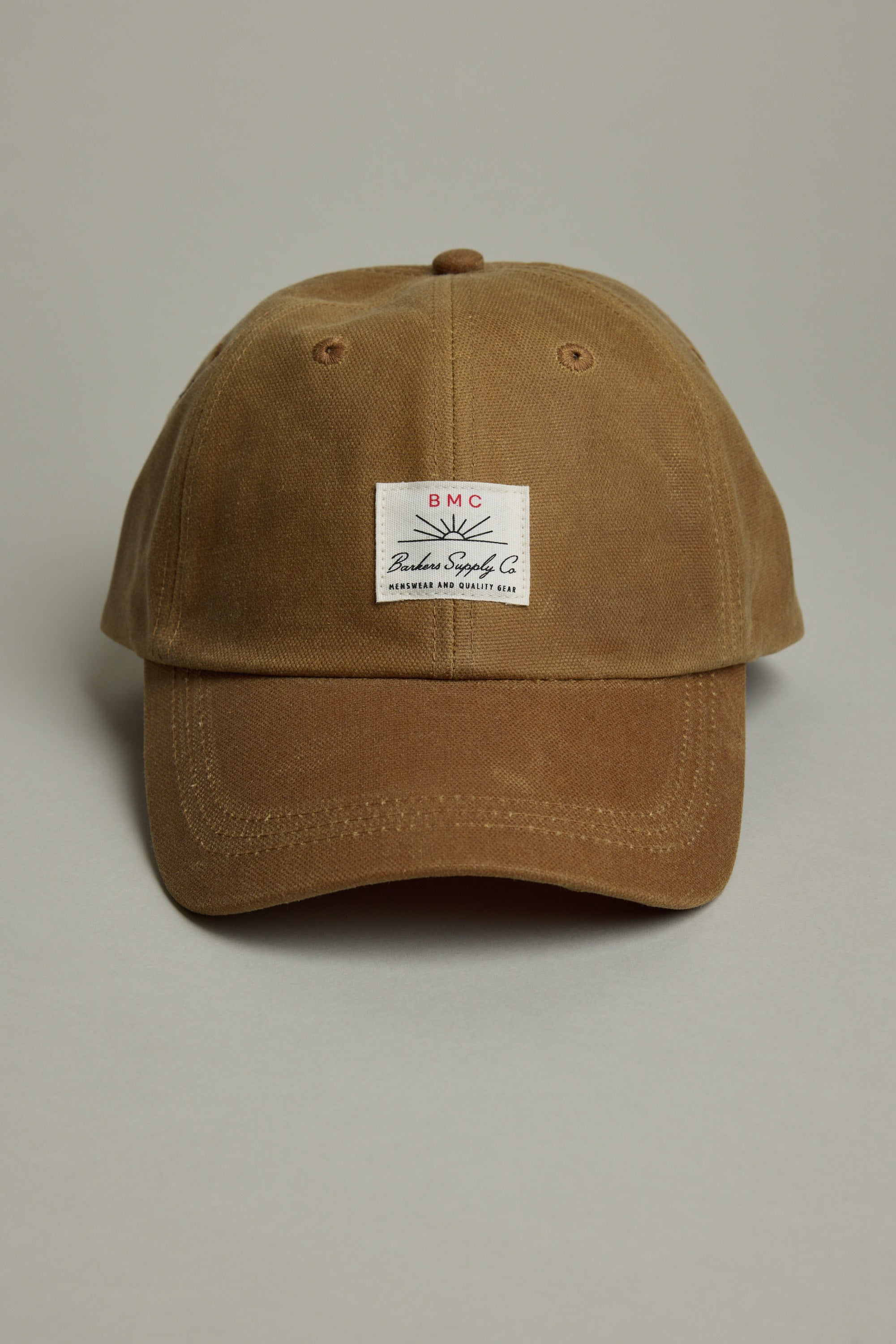 The Barkers Benson Waxed Cotton Cap features brown waxed cotton, a curved brim, and a rectangular white patch with text and graphic. Adjustable for comfort, its heritage style stands out against a neutral backdrop.