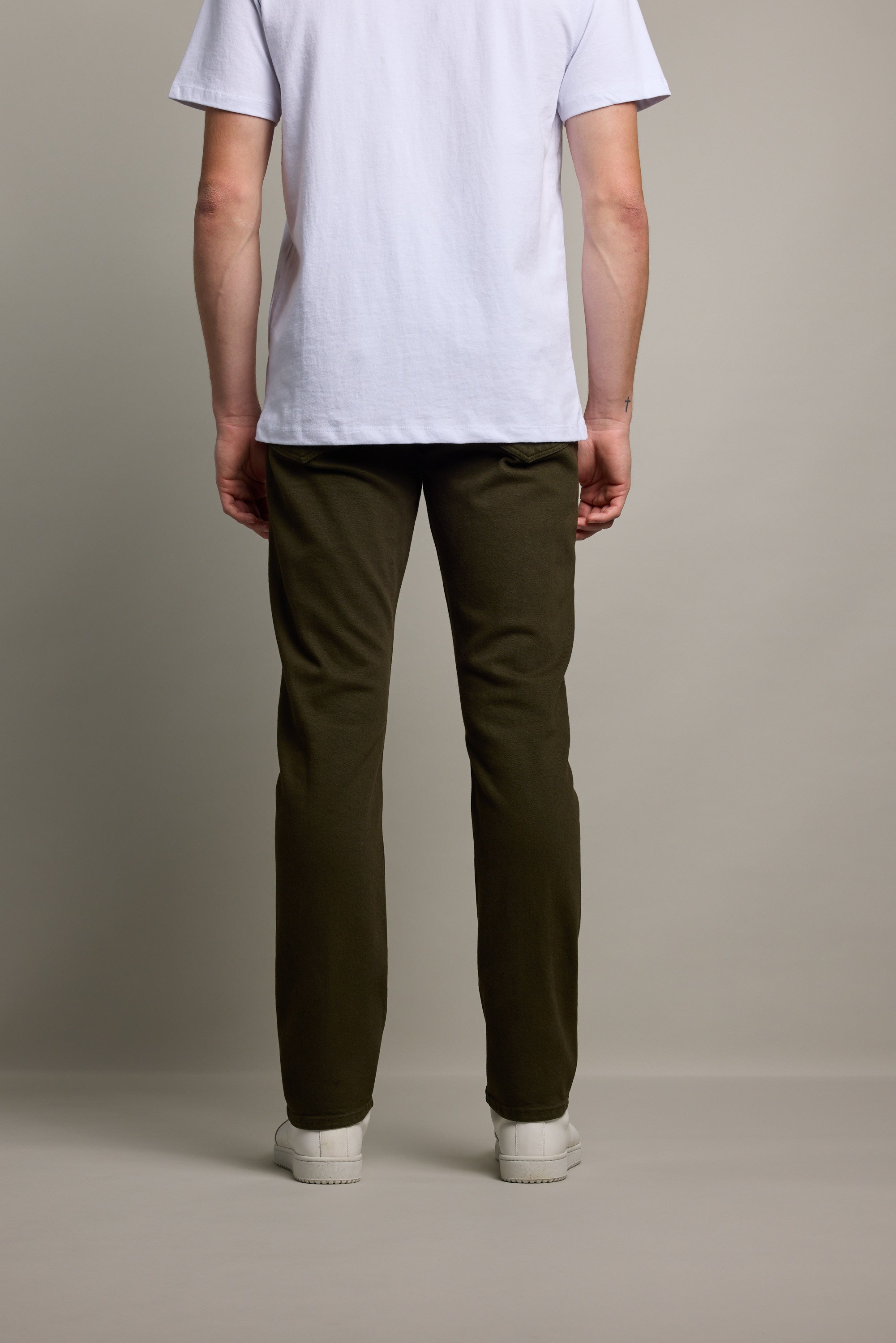 A person stands facing away from the camera, wearing a white short-sleeve t-shirt and Barkers Rambler Slim Coloured Jeans, paired with white sneakers against a plain light-colored background.