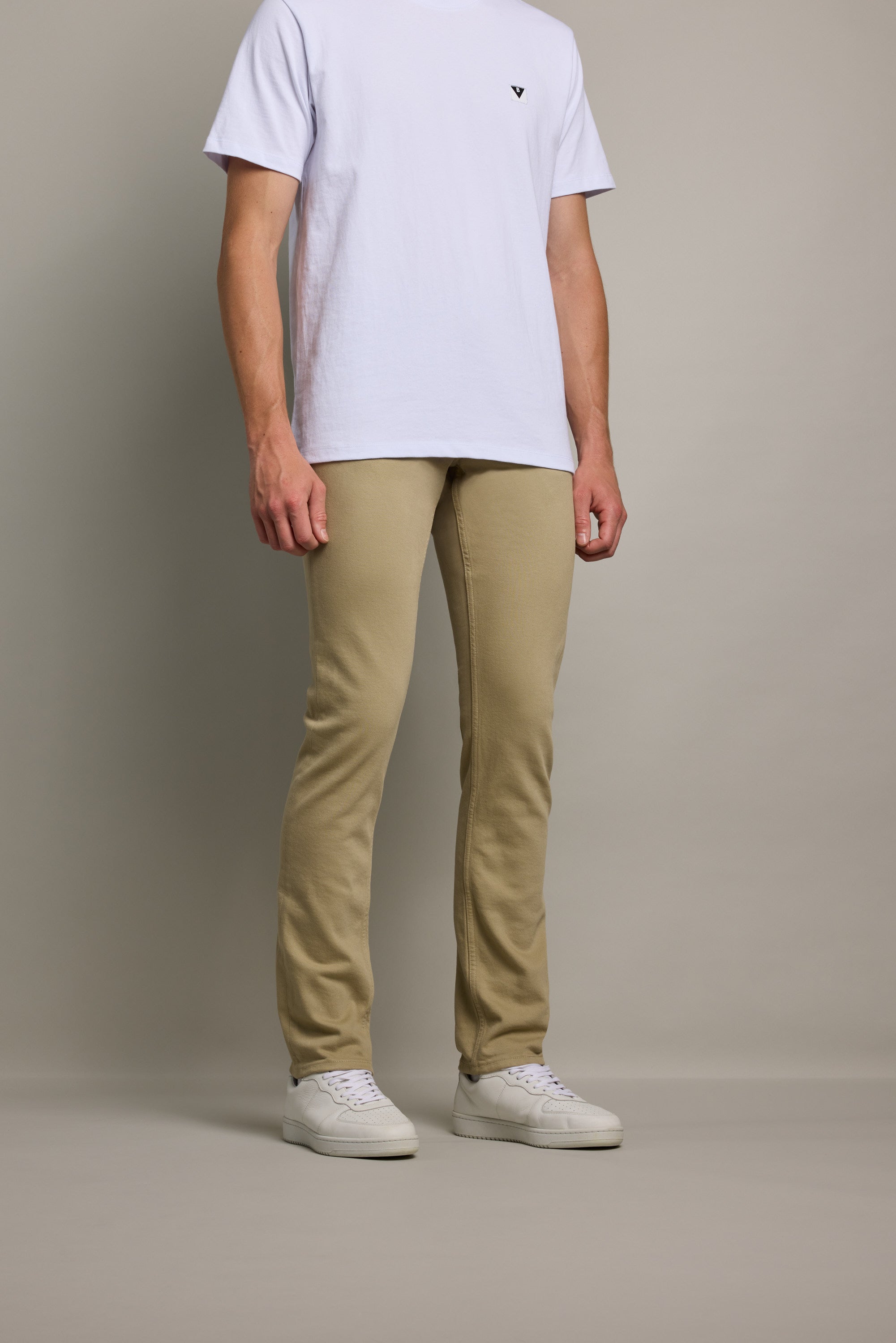 A person wearing Barkers' Rambler Slim Coloured Jean in tan, a plain white T-shirt, and white sneakers stands against a neutral background. The image is cropped from the shoulders down.
