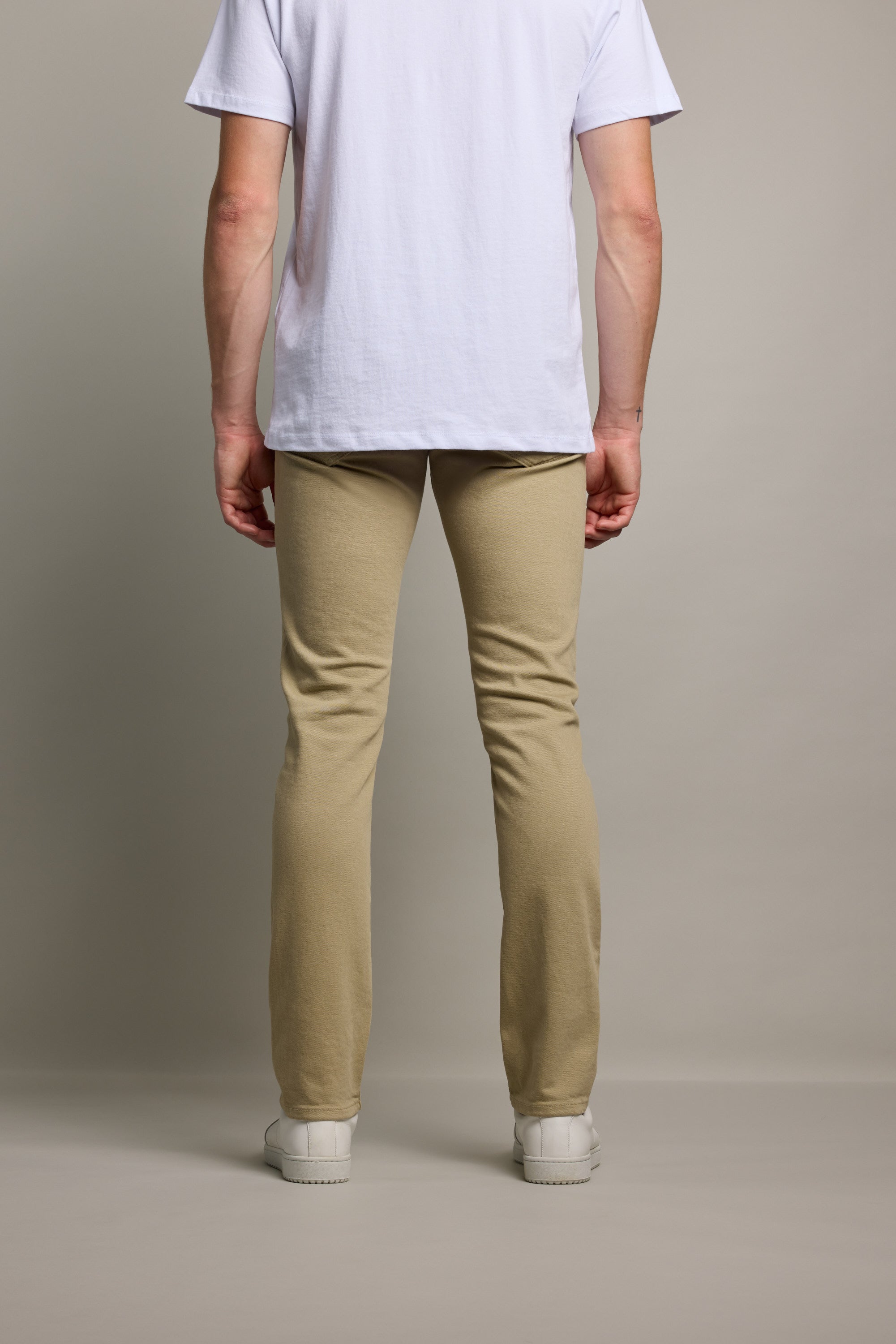 A person stands facing away from the camera, wearing a white T-shirt, Barkers Rambler Slim Coloured Jean, and white sneakers against a plain, light background.