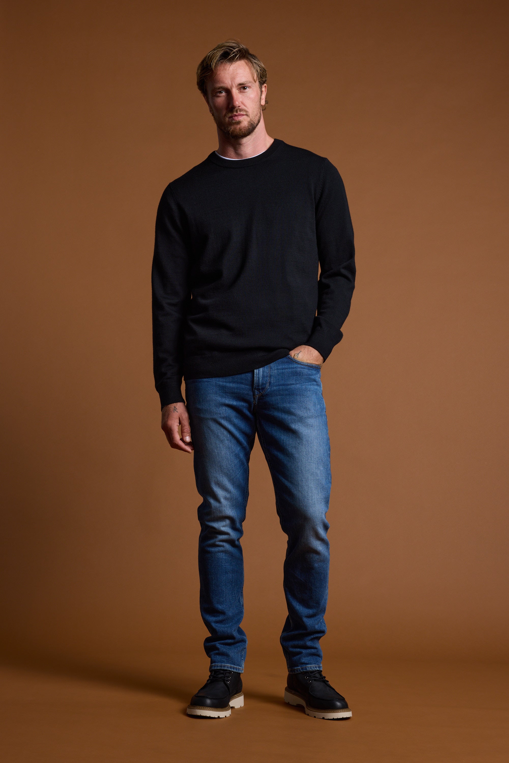 A man with short light hair wears a black Barkers Skyland ZQrx Merino Crew, made from sustainable New Zealand merino wool, paired with blue jeans and black shoes, standing against a plain brown background with one hand in his pocket.