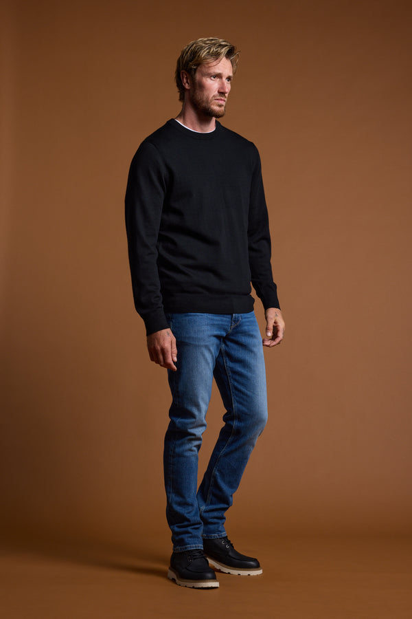 A man with short blond hair and a beard stands against a brown background, wearing the Barkers Skyland ZQrx Merino Crew sweater, blue jeans, and black shoes. He looks to the side with a neutral expression, showcasing the sustainable merino knit.