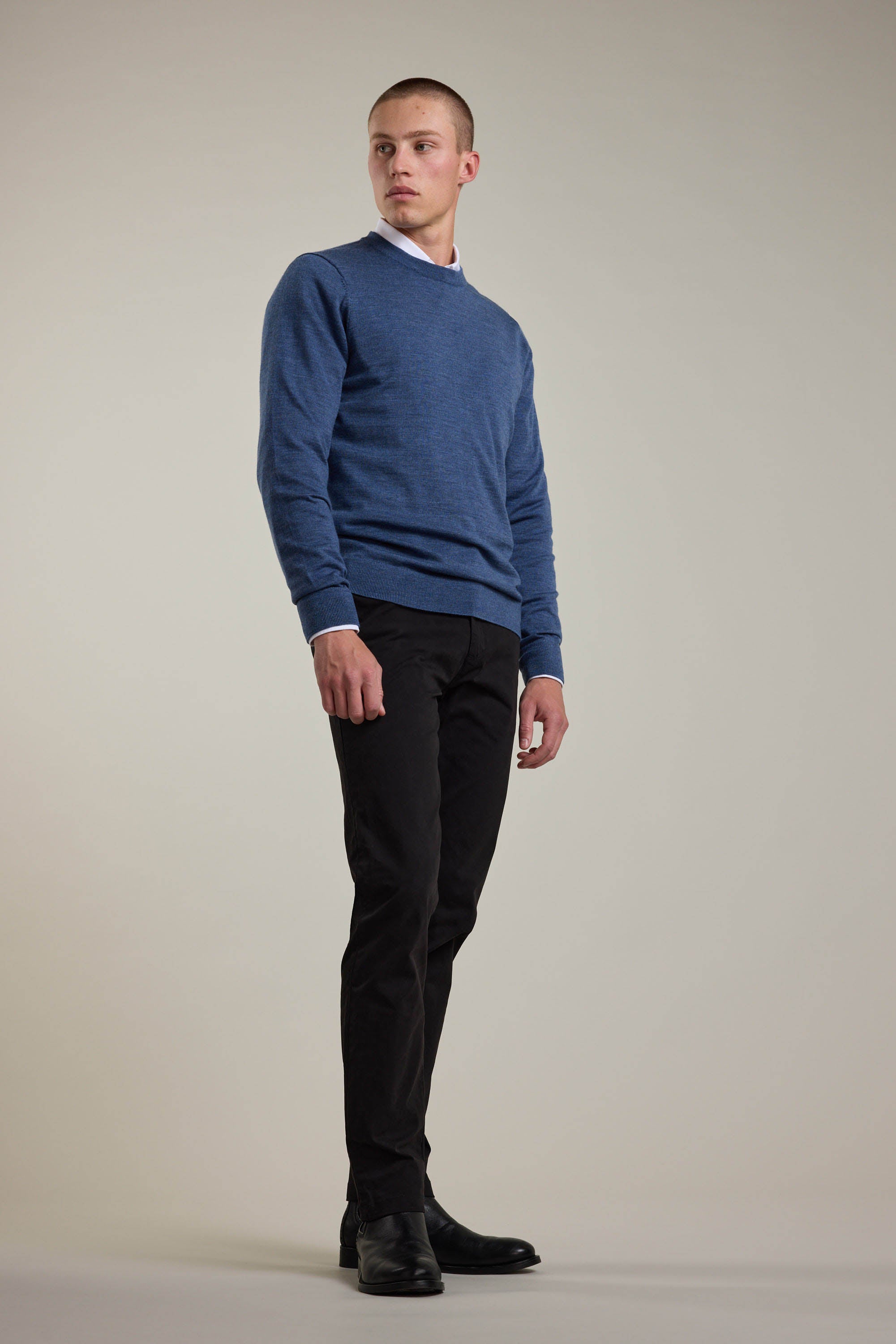 A man stands against a light gray background wearing a blue Barkers Skyland ZQrx Merino Crew sweater made from ethically sourced New Zealand merino wool, paired with black pants and shoes, gazing slightly to the side with a neutral expression.