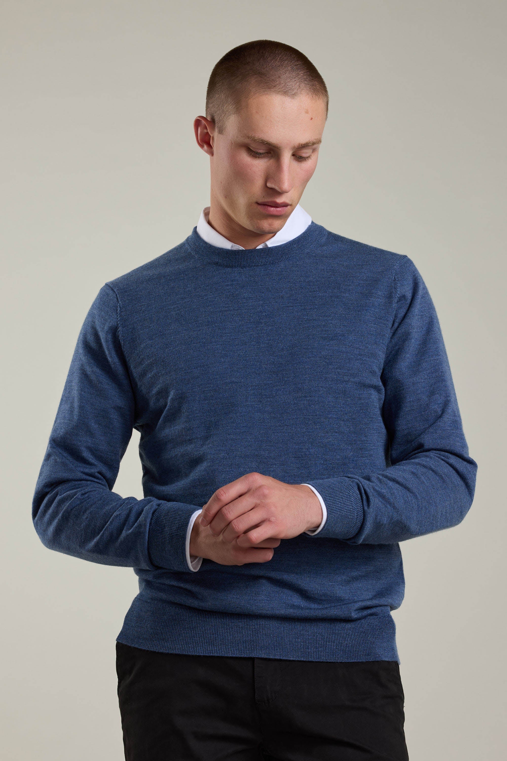 A person wearing a white collared shirt under a Barkers Skyland ZQrx Merino Crew blue sweater and black pants stands against a plain background, looking down with hands clasped in front.