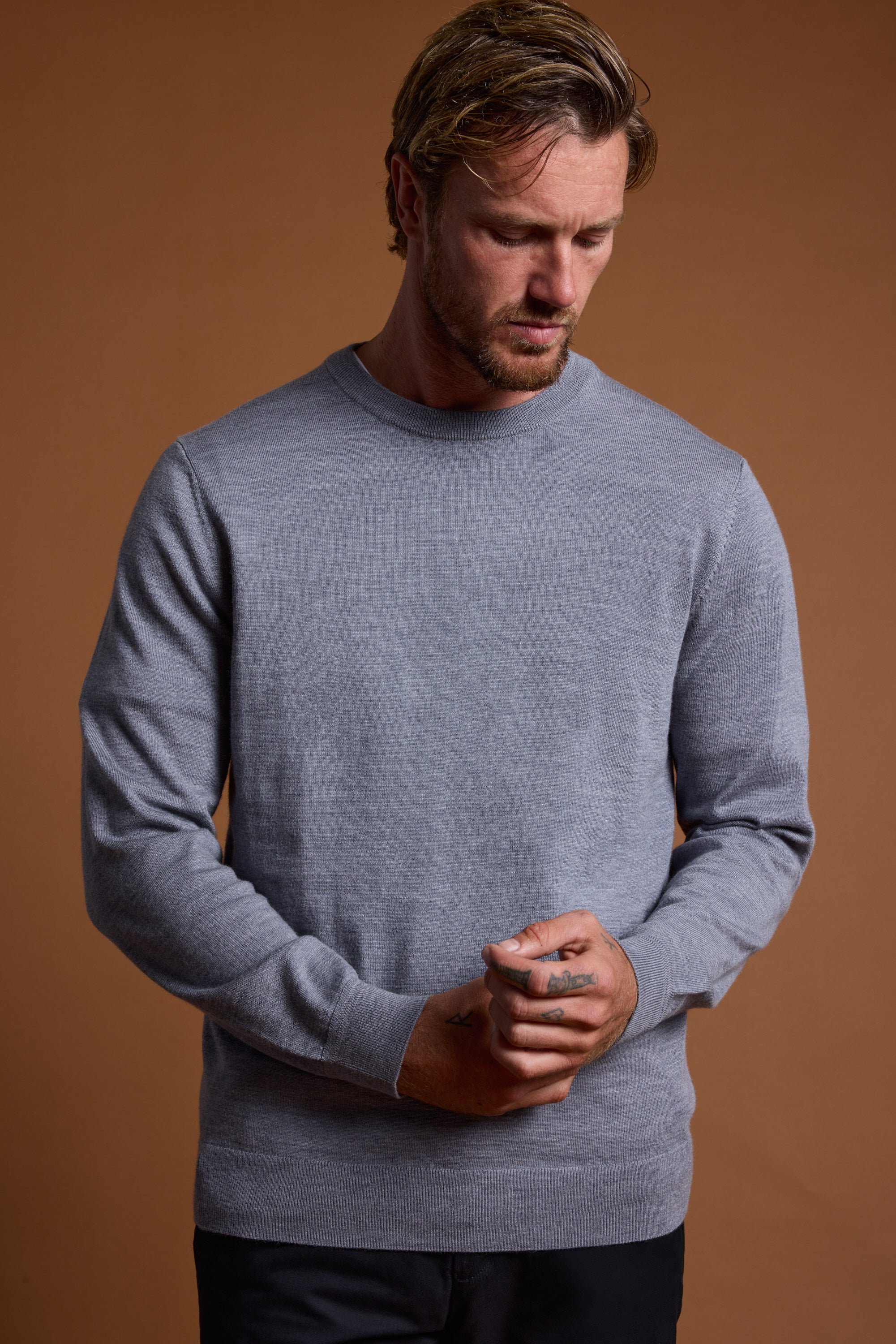 A man with short, light brown hair and a beard wears the Barkers Skyland ZQrx Merino Crew, made from sustainable New Zealand merino wool. He stands against a brown background, looking down with his hands clasped.