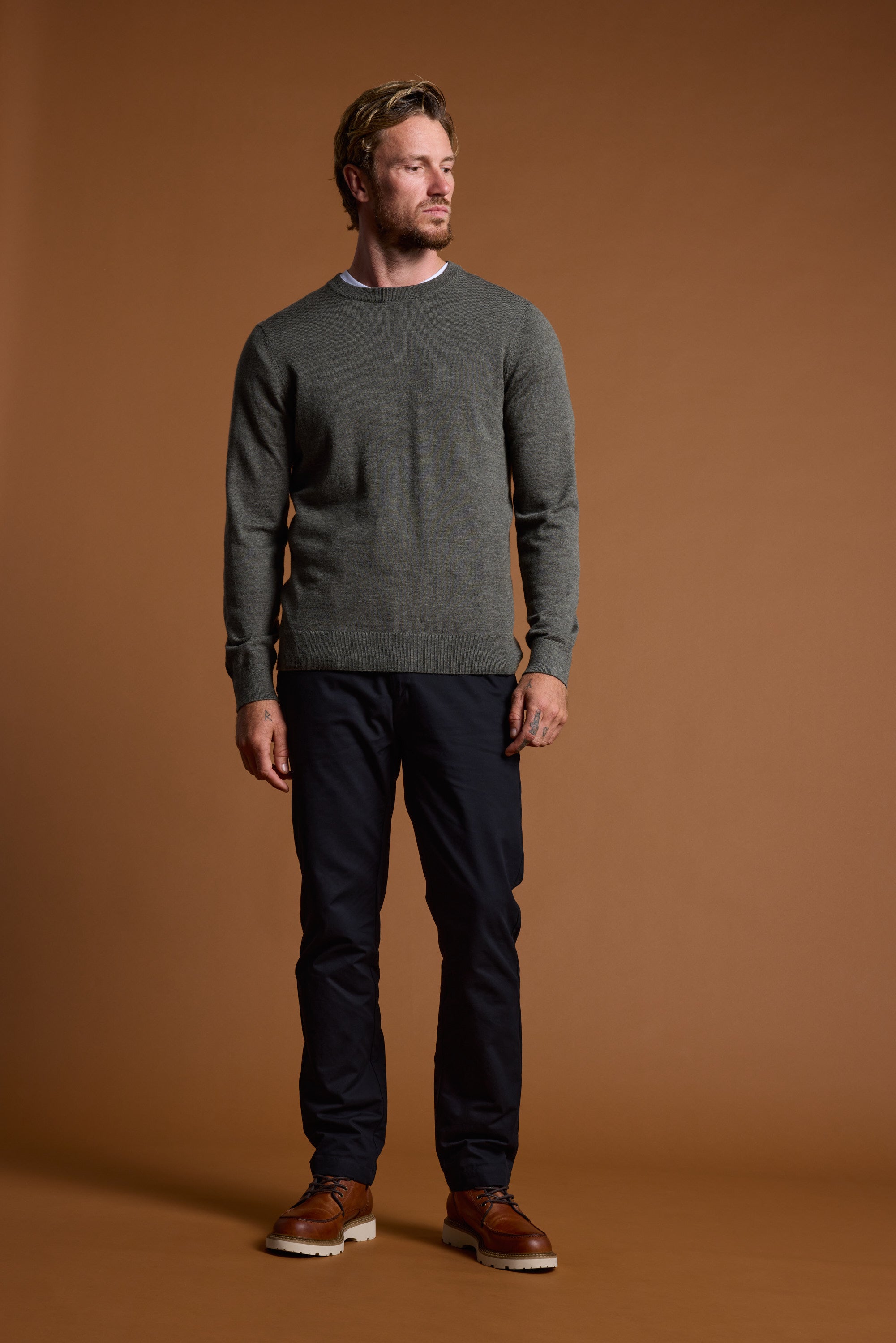 A man with short hair and a beard stands against a brown background, wearing the Barkers Skyland ZQrx Merino Crew made from ethically sourced New Zealand merino wool. He looks left with a neutral expression, paired with dark pants and brown shoes.