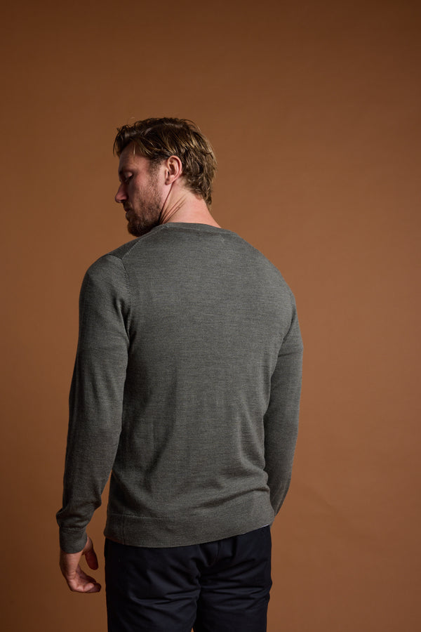 A man with light brown hair, wearing a Barkers Skyland ZQrx Merino Crew in gray and dark pants, stands facing away from the camera against a plain brown background.