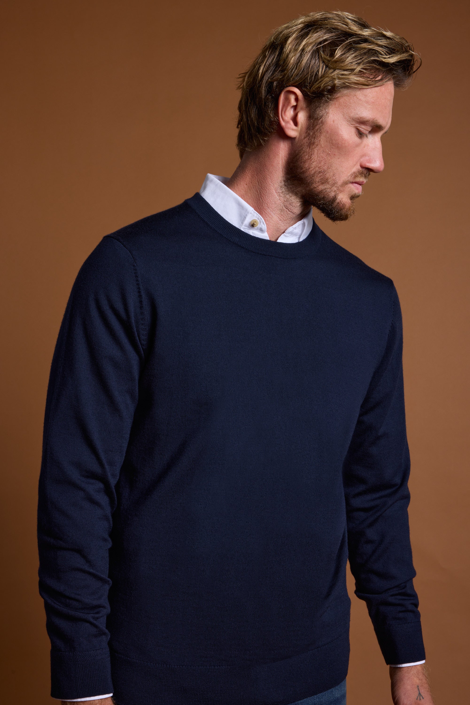 A man with blond hair and a short beard wears a navy blue Barkers Skyland ZQrx Merino Crew over a white collared shirt, looking down against a brown background. This sustainable sweater is made from premium New Zealand merino wool.