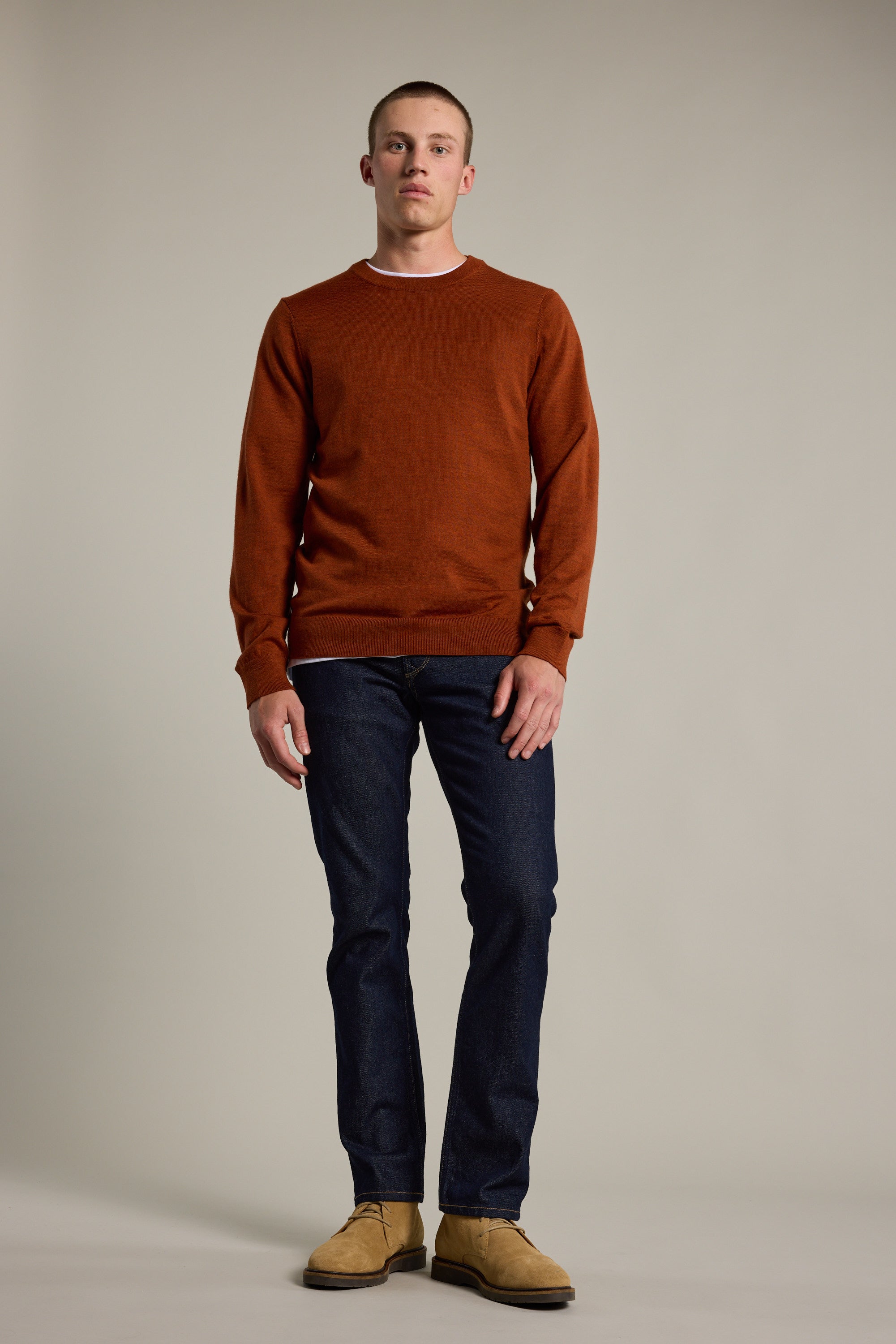 A man stands facing forward, wearing the Barkers Skyland ZQrx Merino Crew in burnt orange, paired with dark blue jeans and tan suede shoes, set against a plain light gray background.