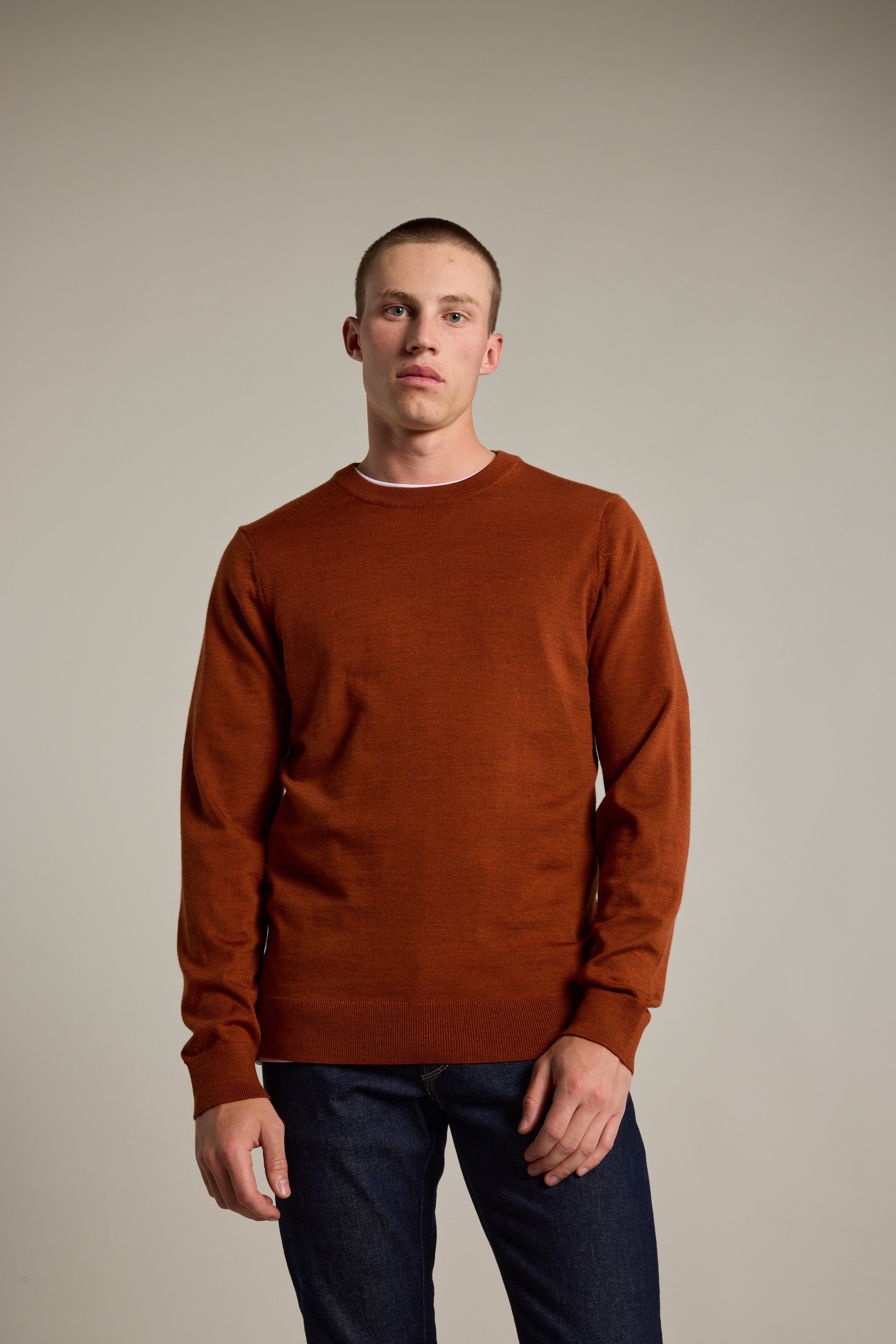 A young man with short hair stands against a beige background, wearing a rust-colored Barkers Skyland ZQrx Merino Crew and dark blue jeans. He has a neutral expression, his left hand resting slightly in his pocket.