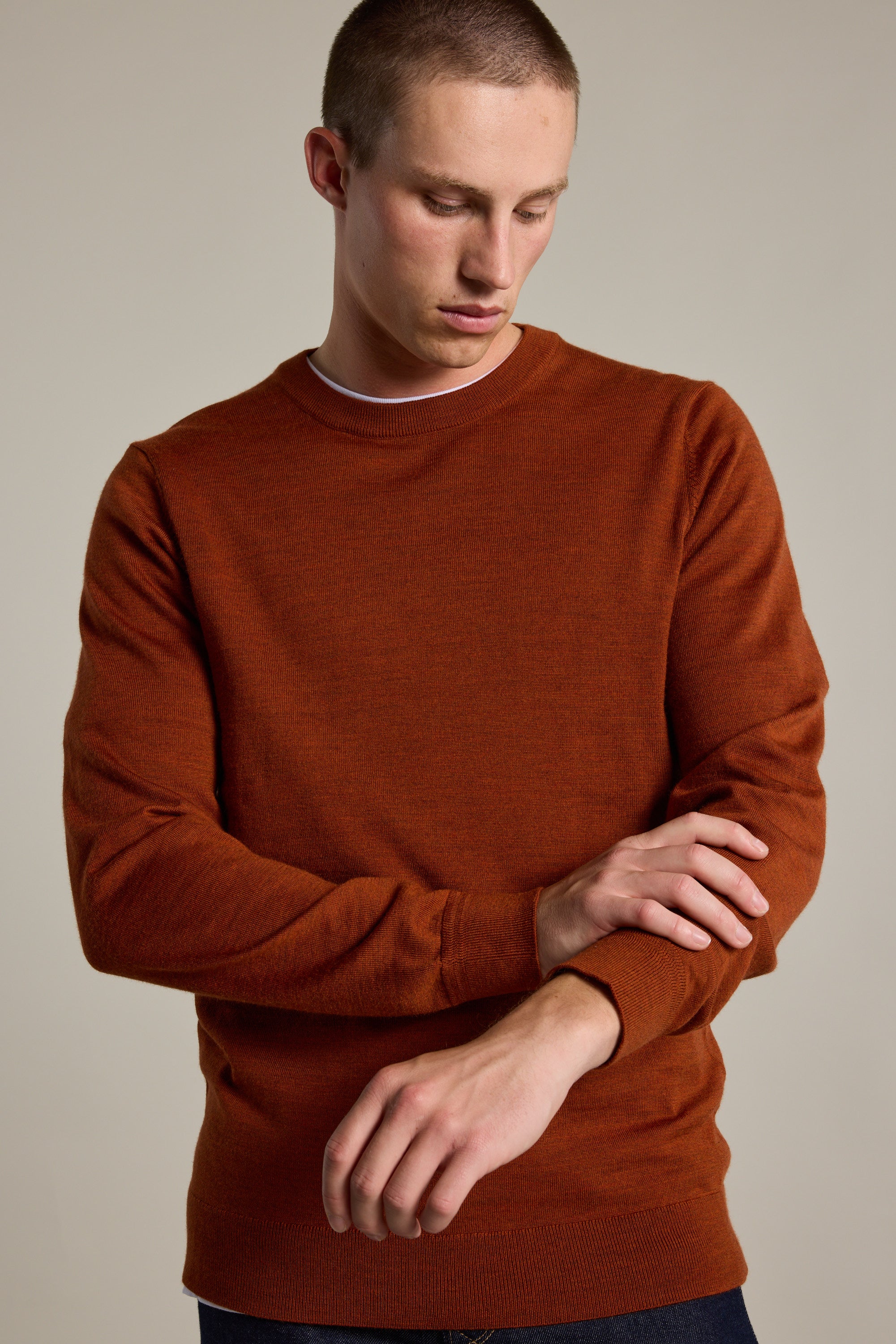A man wearing the Barkers Skyland ZQrx Merino Crew, a rust-colored sustainable sweater, adjusts his sleeve while standing against a plain, light background.